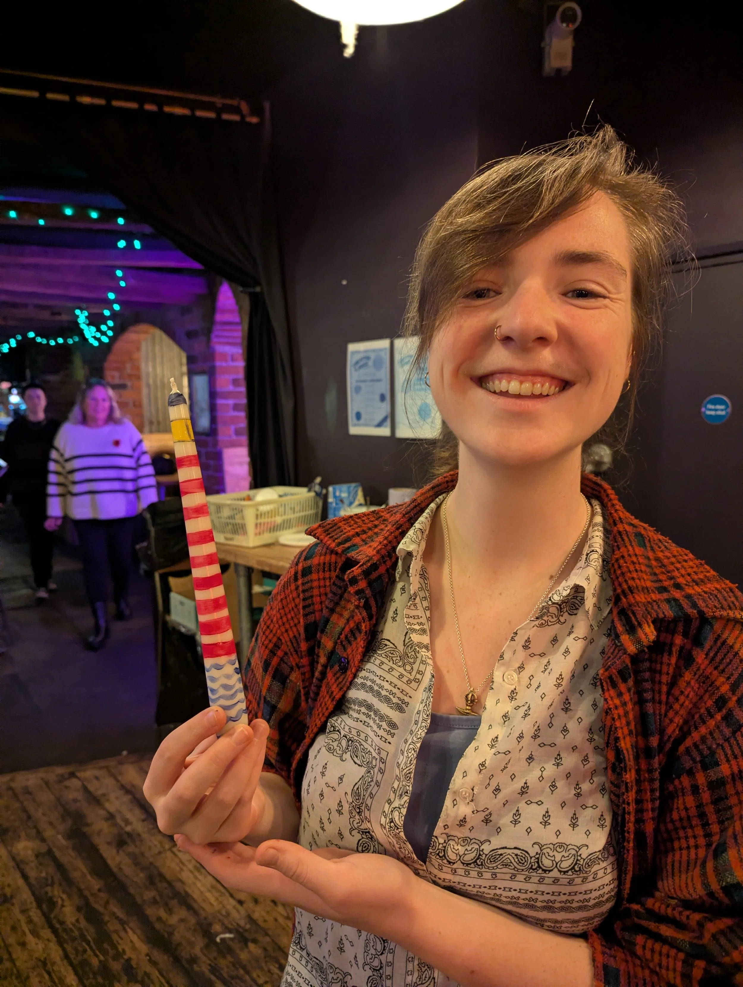 A young woman with short hair, wearing a patterned shirt and a red plaid jacket, smiling and holding a candle with red, white, and blue stripes inside an indoor venue with exposed brick walls and other people in the background.