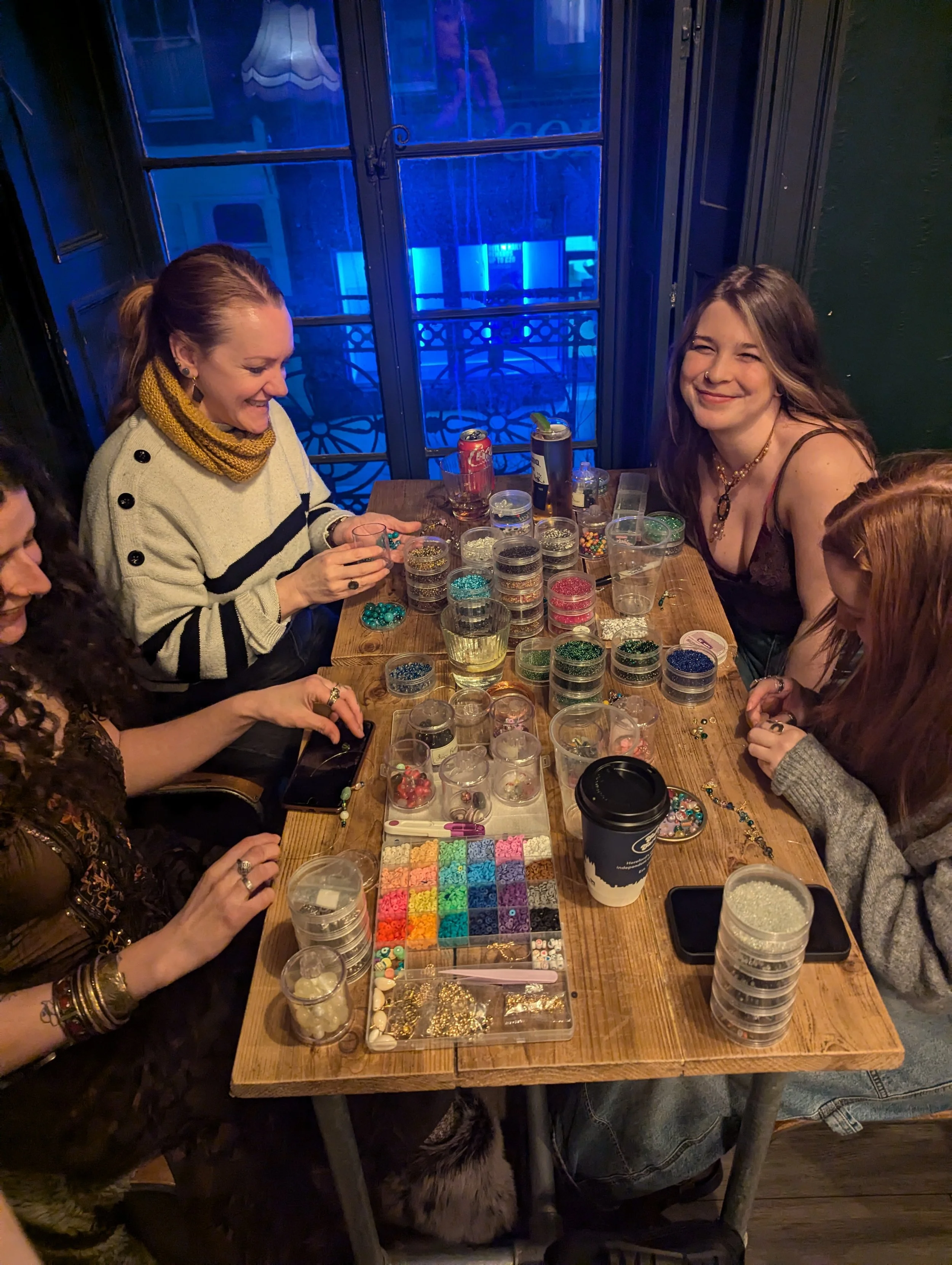 Group of women sitting around a table filled with containers of colorful beads and jewelry-making supplies, happily working on craft projects in a cozy, dimly lit room.