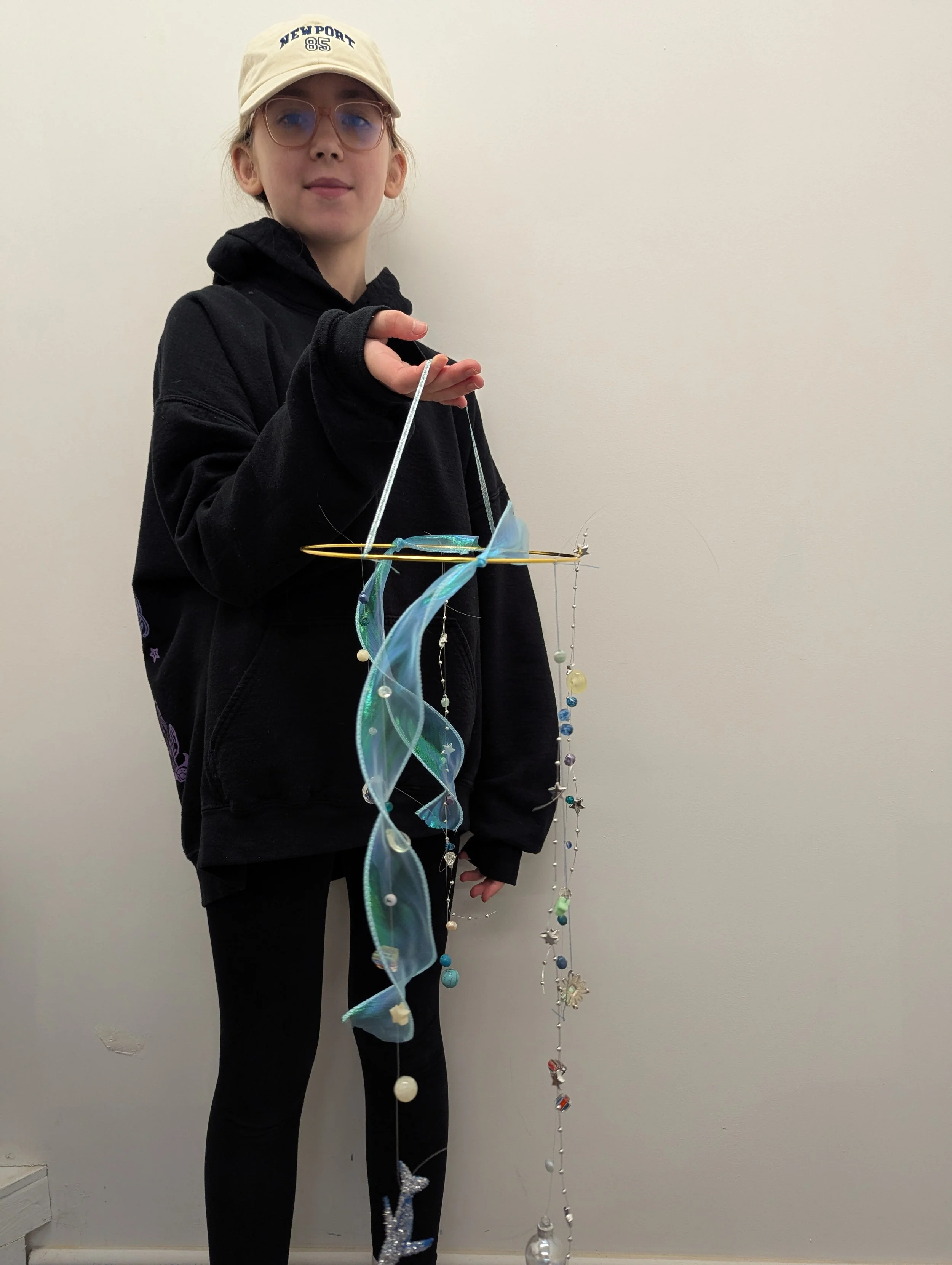A young girl wearing glasses, a beige cap, and a black hoodie, holding a mobile hanging mobile with blue and clear decorations against a plain white wall.