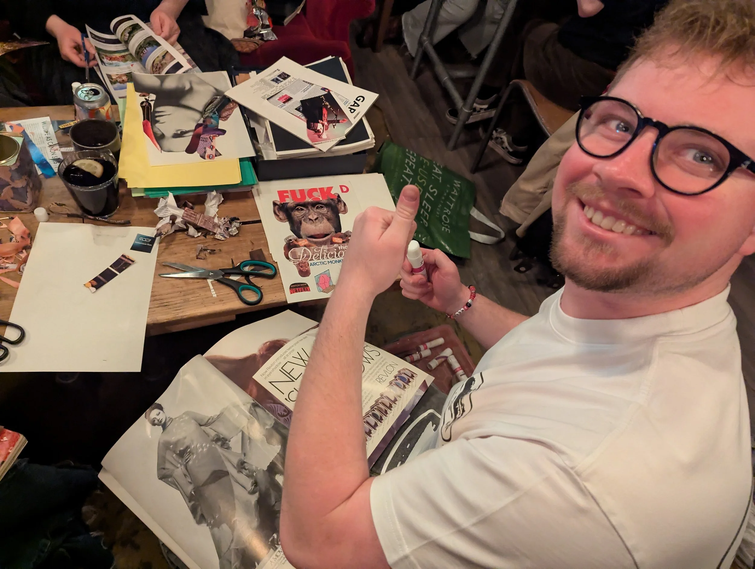 A man smiling at the camera, seated at a cluttered table with various papers, magazines, scissors, and art supplies, holding a glue stick.