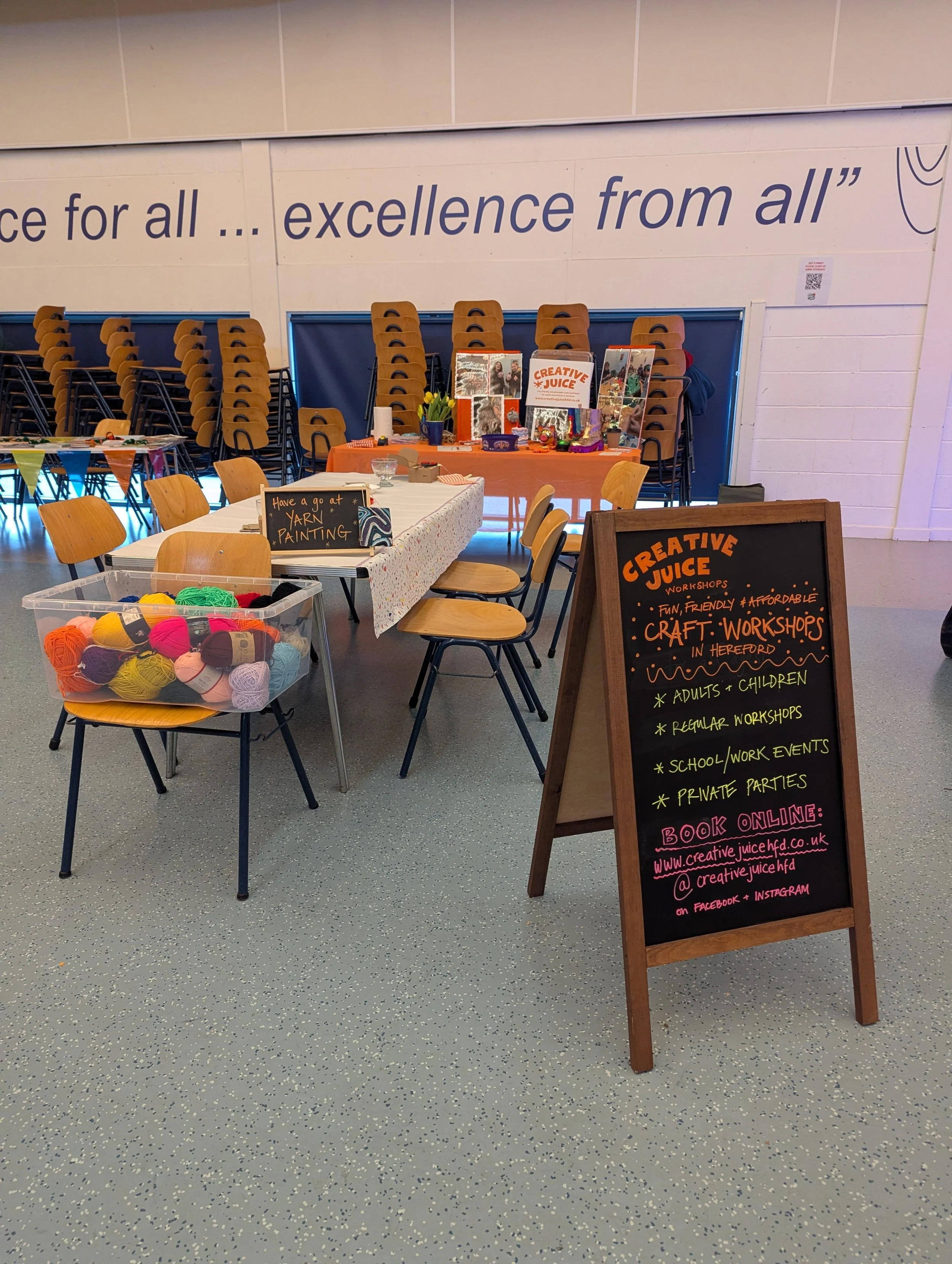 An indoor craft event space with a chalkboard sign advertising Creative Juice workshops for adults, children, school, work events, and private parties, with information about booking online on a black background. There are tables with yarn balls, cha