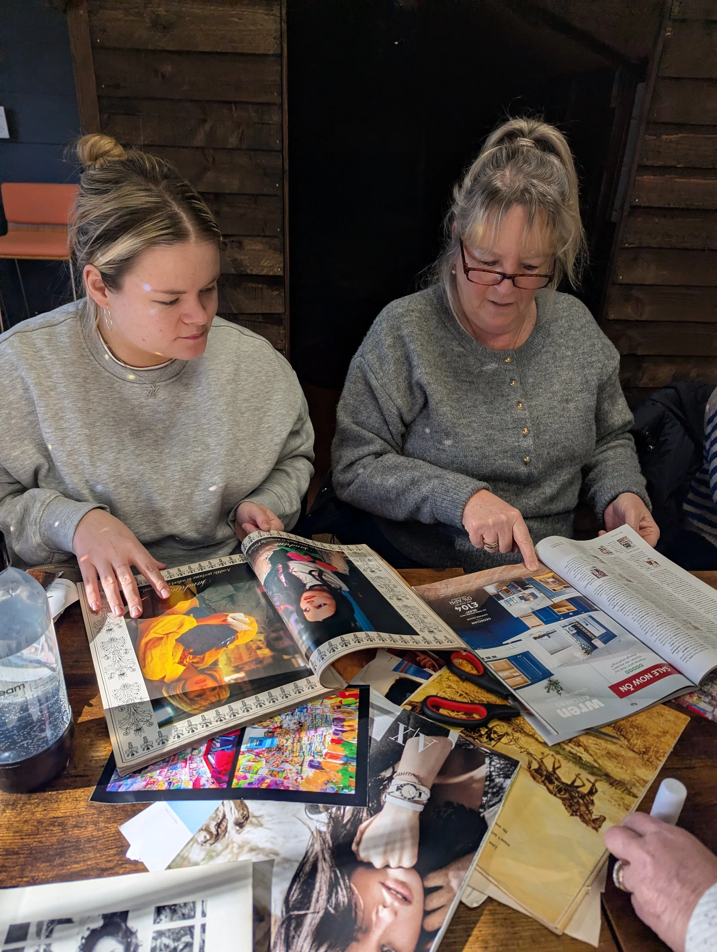 Two women sitting at a wooden table looking through magazines, surrounded by scissors, a glue stick, and other craft supplies.