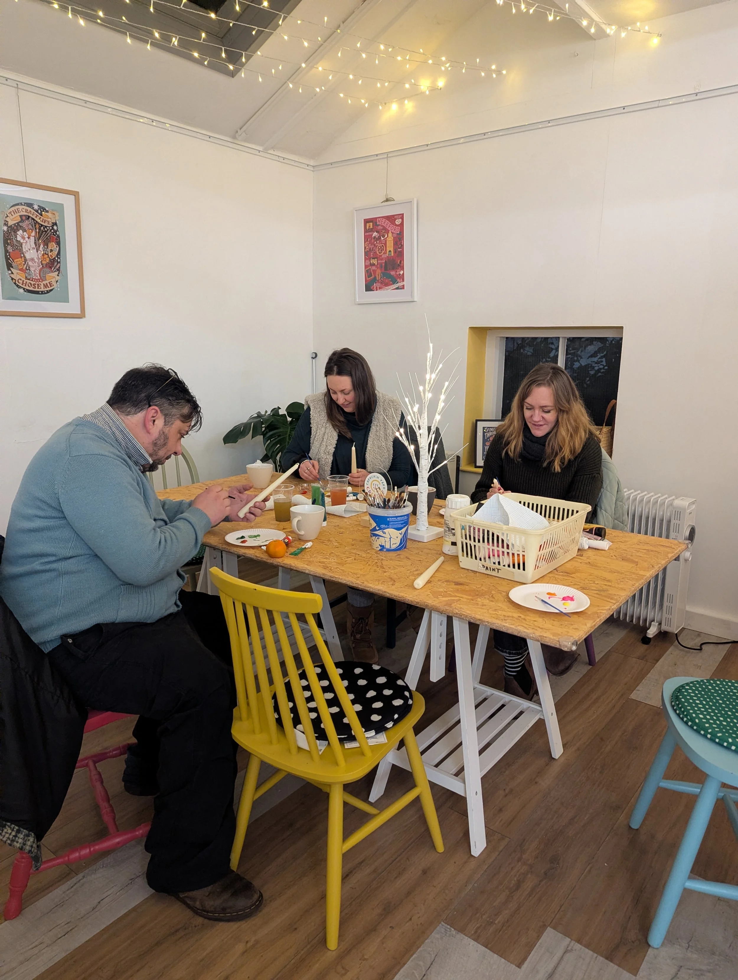 Three people sitting at a wooden table decorating pottery, with art supplies and a small white decorative tree with lights on the table, in a room with white walls, string lights, and framed art.