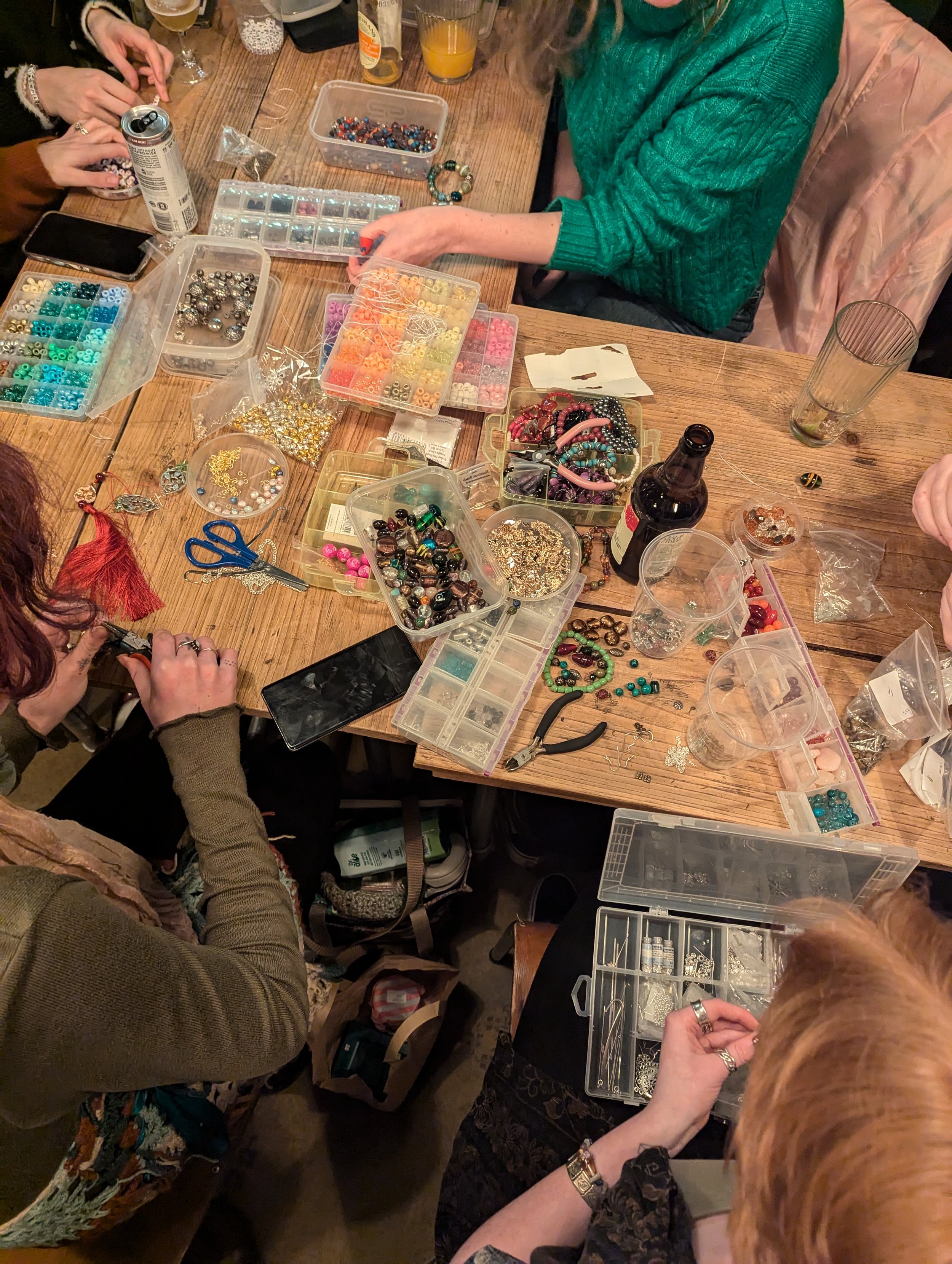 People gathered around a wooden table making jewelry with various beads, tools, and jewelry supplies spread across the table.