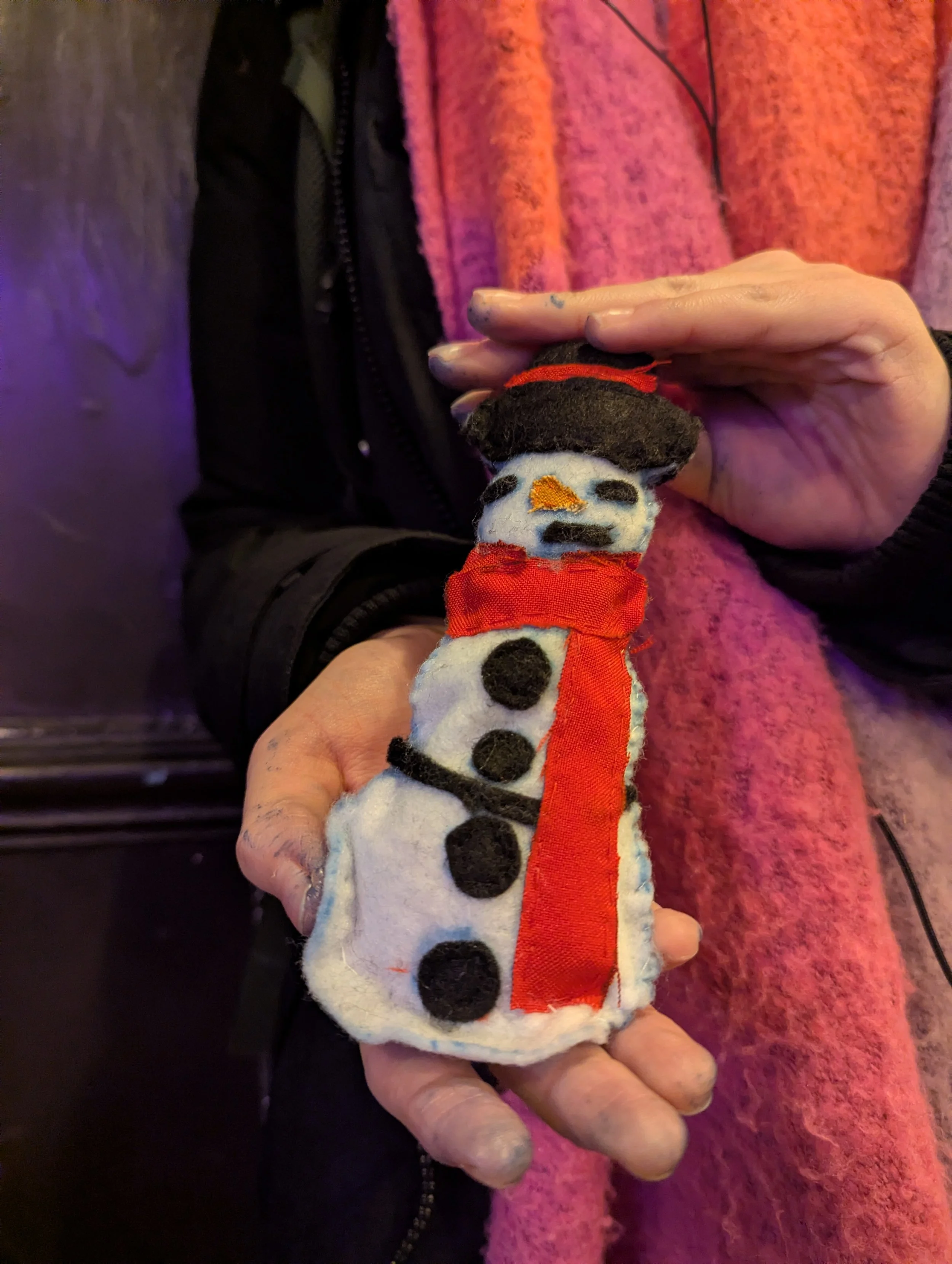 A person holding a small, plush snowman made of fabric with black felt buttons, a black felt hat with a red band, a red scarf, and an orange nose. The person is wearing a pink scarf and a dark jacket.