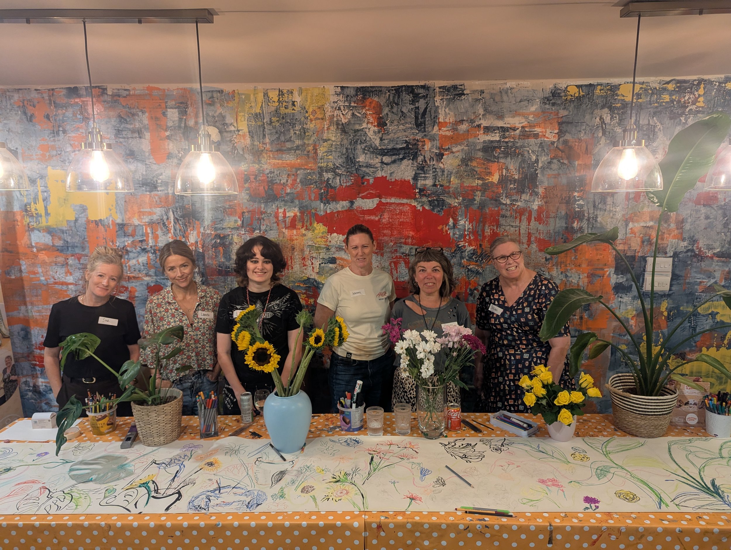 A group of six women standing behind a table covered with colorful drawings and art supplies, in front of a vibrant, abstract wall painting, with hanging lights and a large plant on the right.