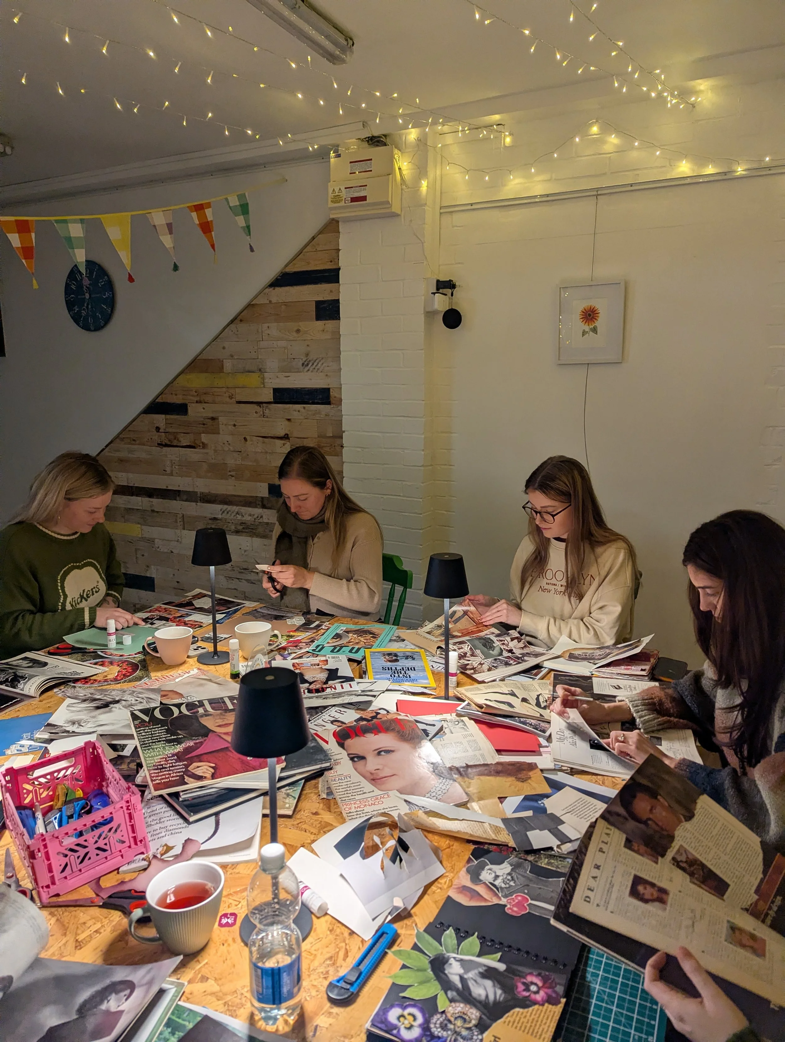 Four women sit around a wooden table covered with magazines, scissors, glue, and arts and crafts supplies, engaged in a crafting activity in a cozy indoor space decorated with string lights, a small ukulele, and colorful bunting.
