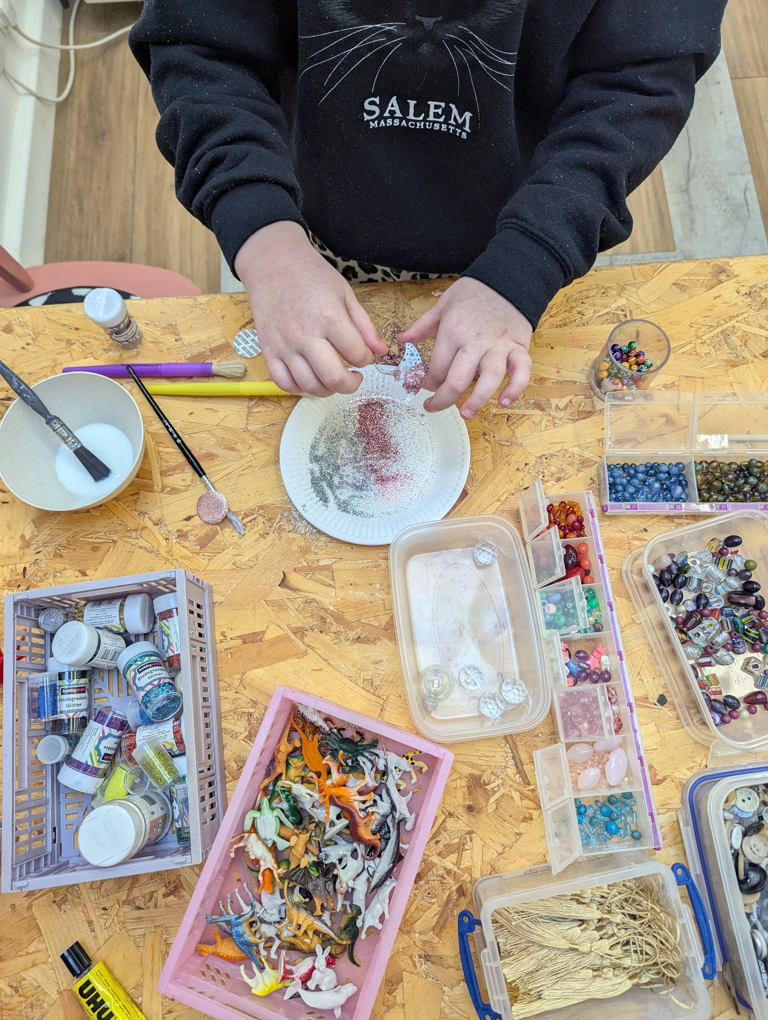 A person crafting with glitter and small decorative items on a wooden table, surrounded by storage containers filled with beads, miniature figures, and craft supplies.