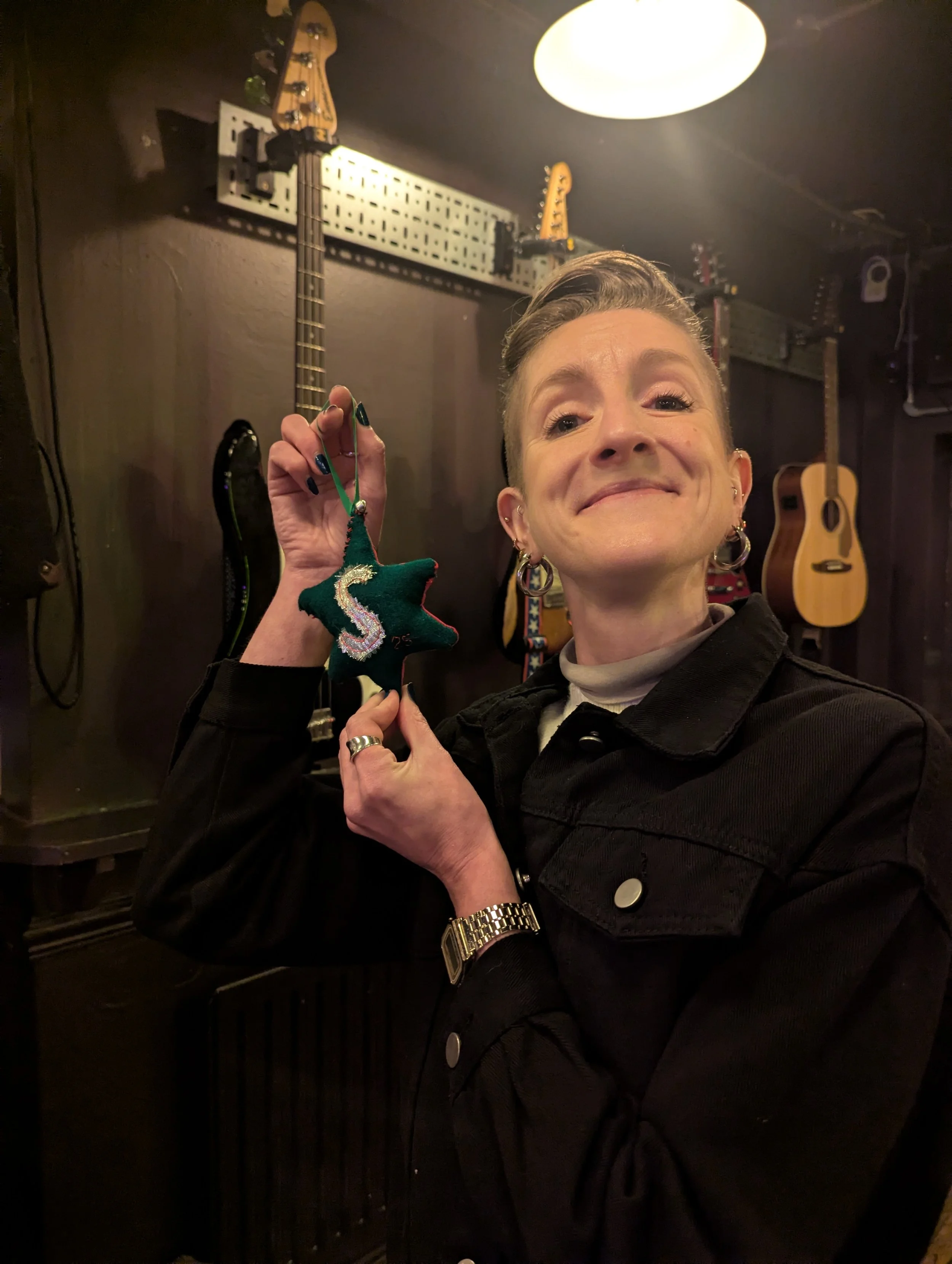 A person smiling and holding a green star-shaped ornament with a glittery letter 'S' on it, in a room with guitars hanging on the wall behind them.