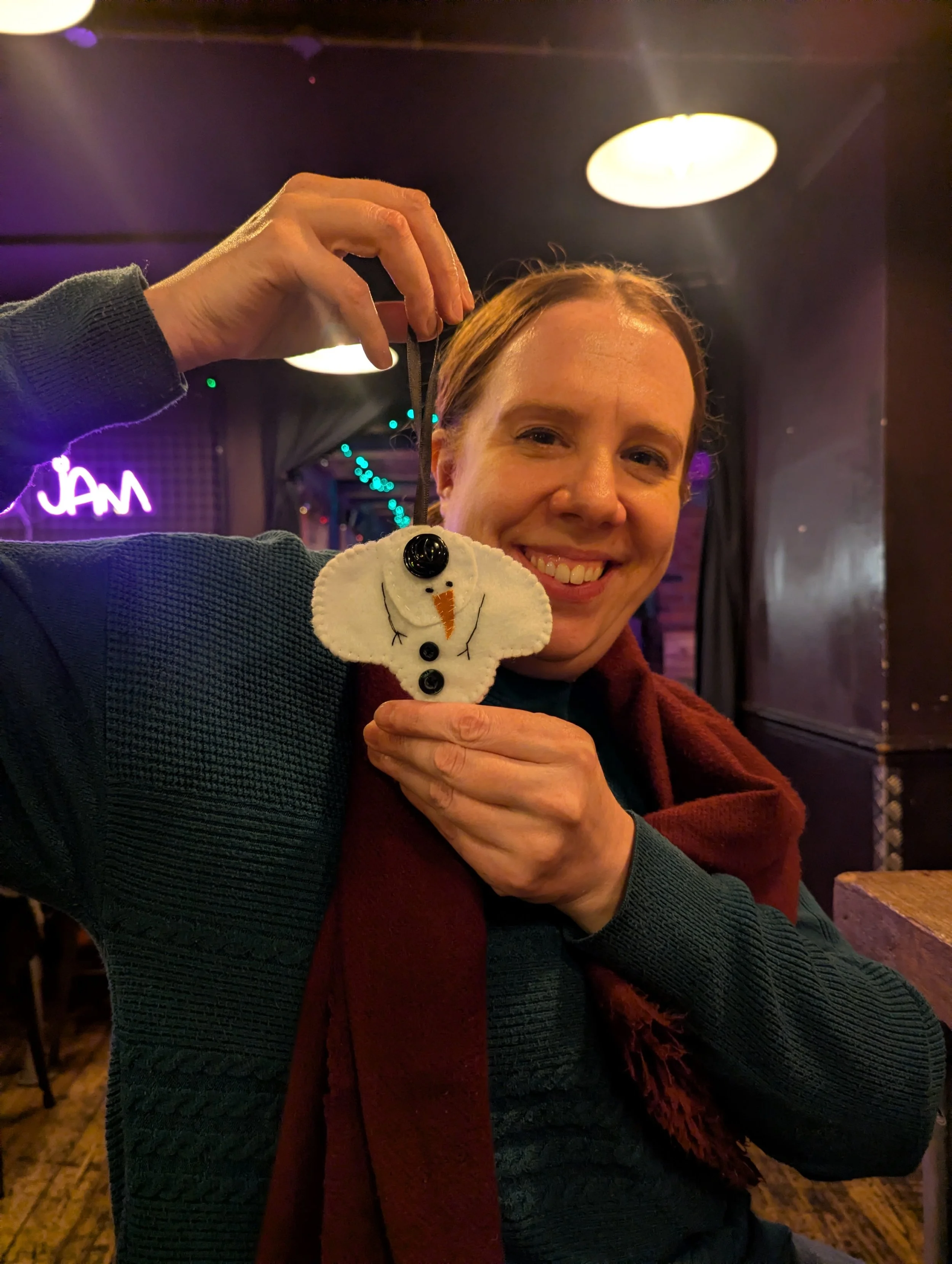 A smiling woman holding a snowman ornament in a dimly lit indoor setting with purple lighting and a neon sign in the background.