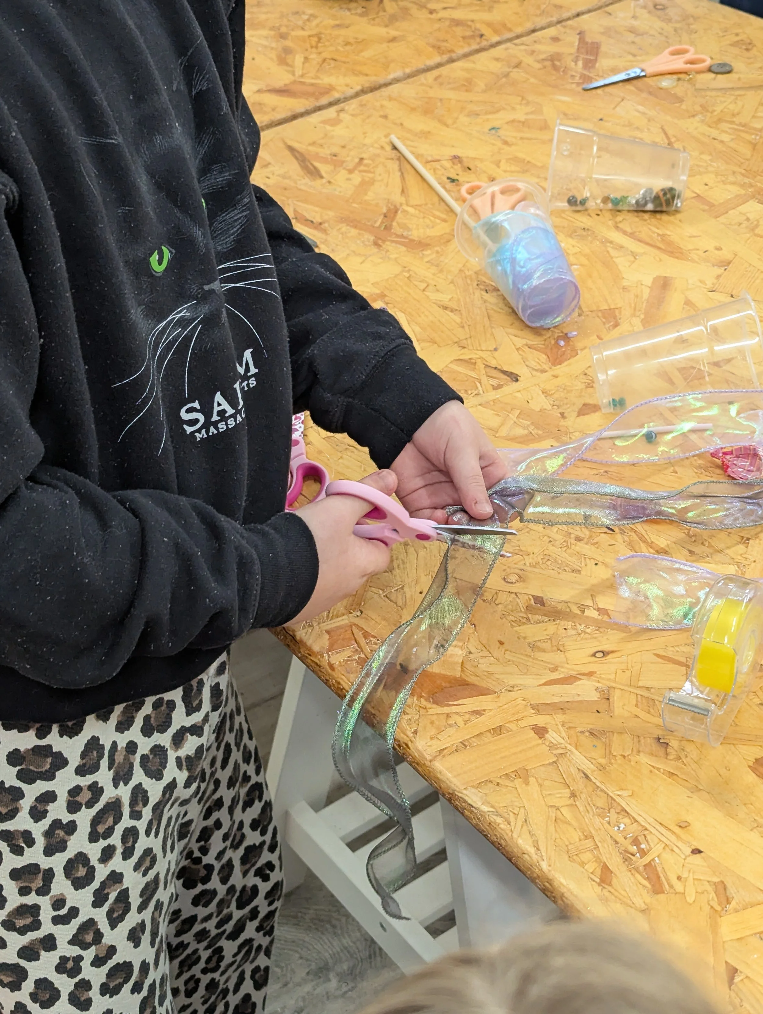 A person cutting a piece of iridescent ribbon with pink scissors on a wooden worktable surrounded by crafting supplies.