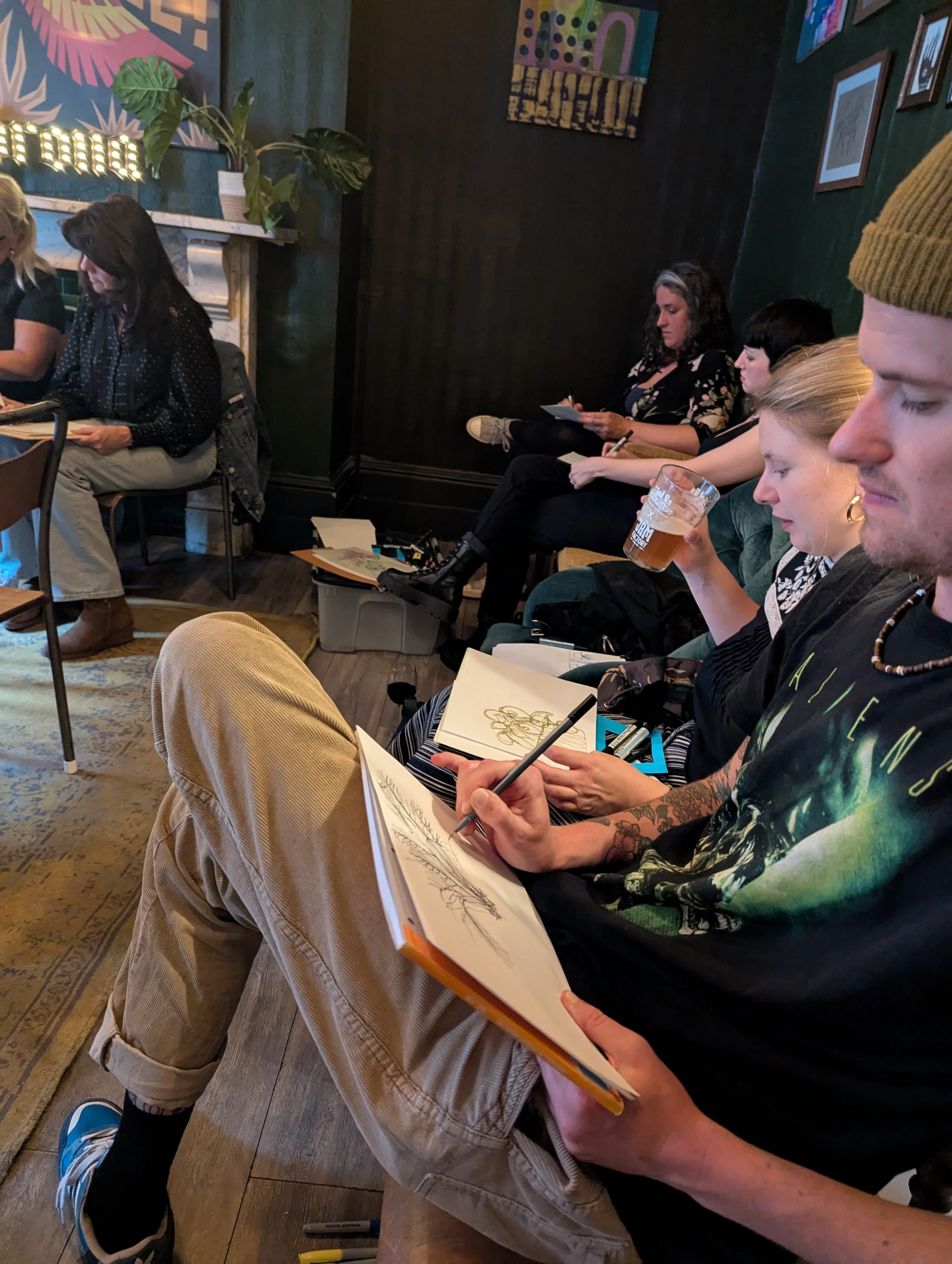 A group of people sitting closely together in a cozy, decorated room, some are sketching in notebooks, one person is holding a glass of beer, others are taking notes, with art on the walls and plants in the background.