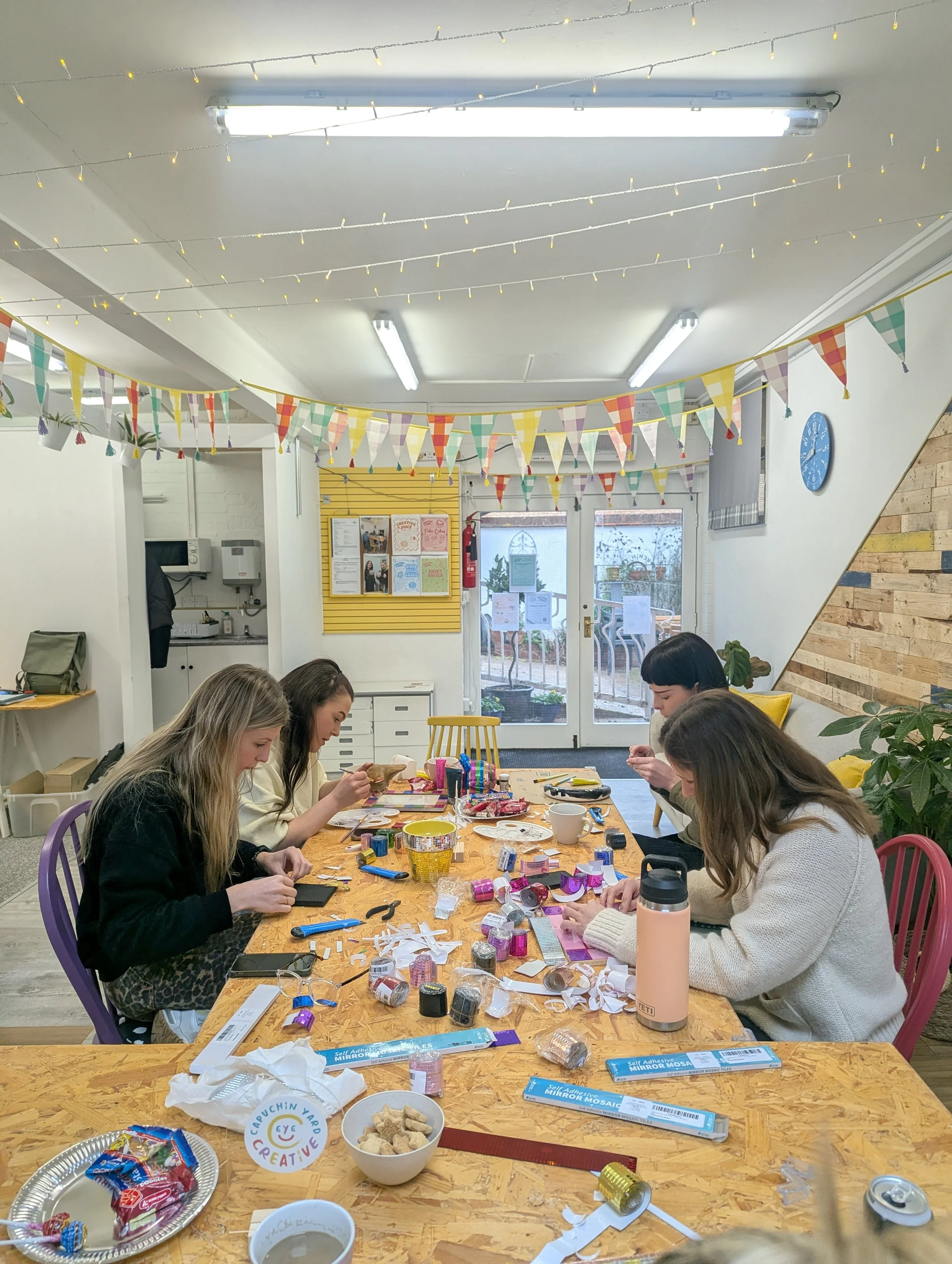 Four women sitting at a table decorating, surrounded by craft supplies under colorful bunting and fairy lights in a decorated room.