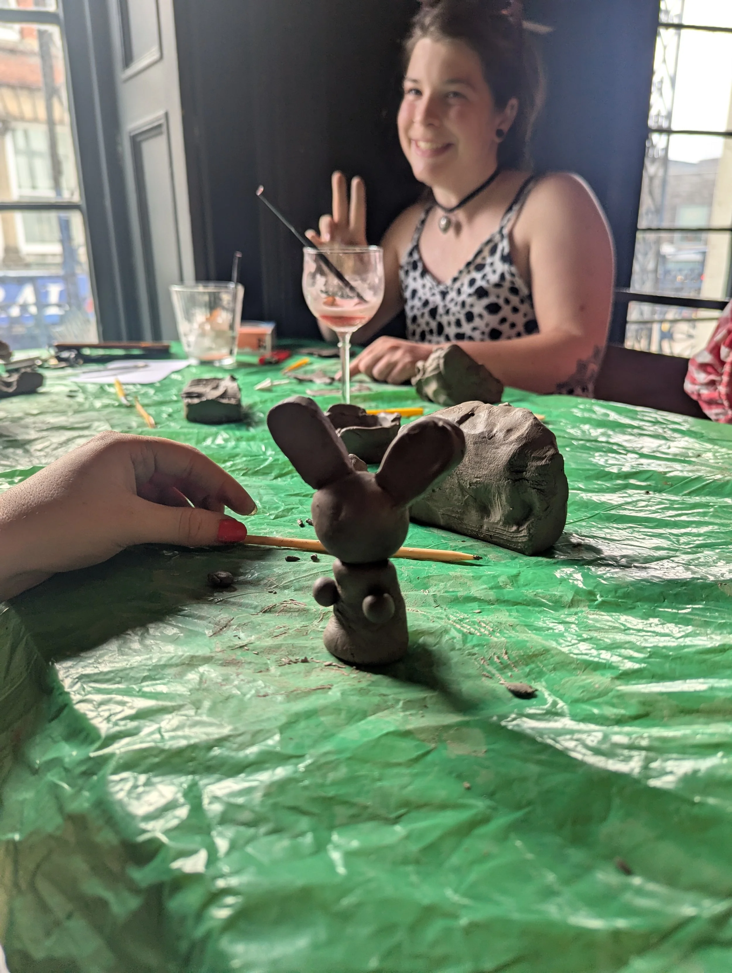 A woman with a tattoo on her arm making a peace sign in a dimly lit room with large windows. A bunny-shaped clay sculpture and other clay projects are on a green plastic-covered table in the foreground, along with art tools and materials.