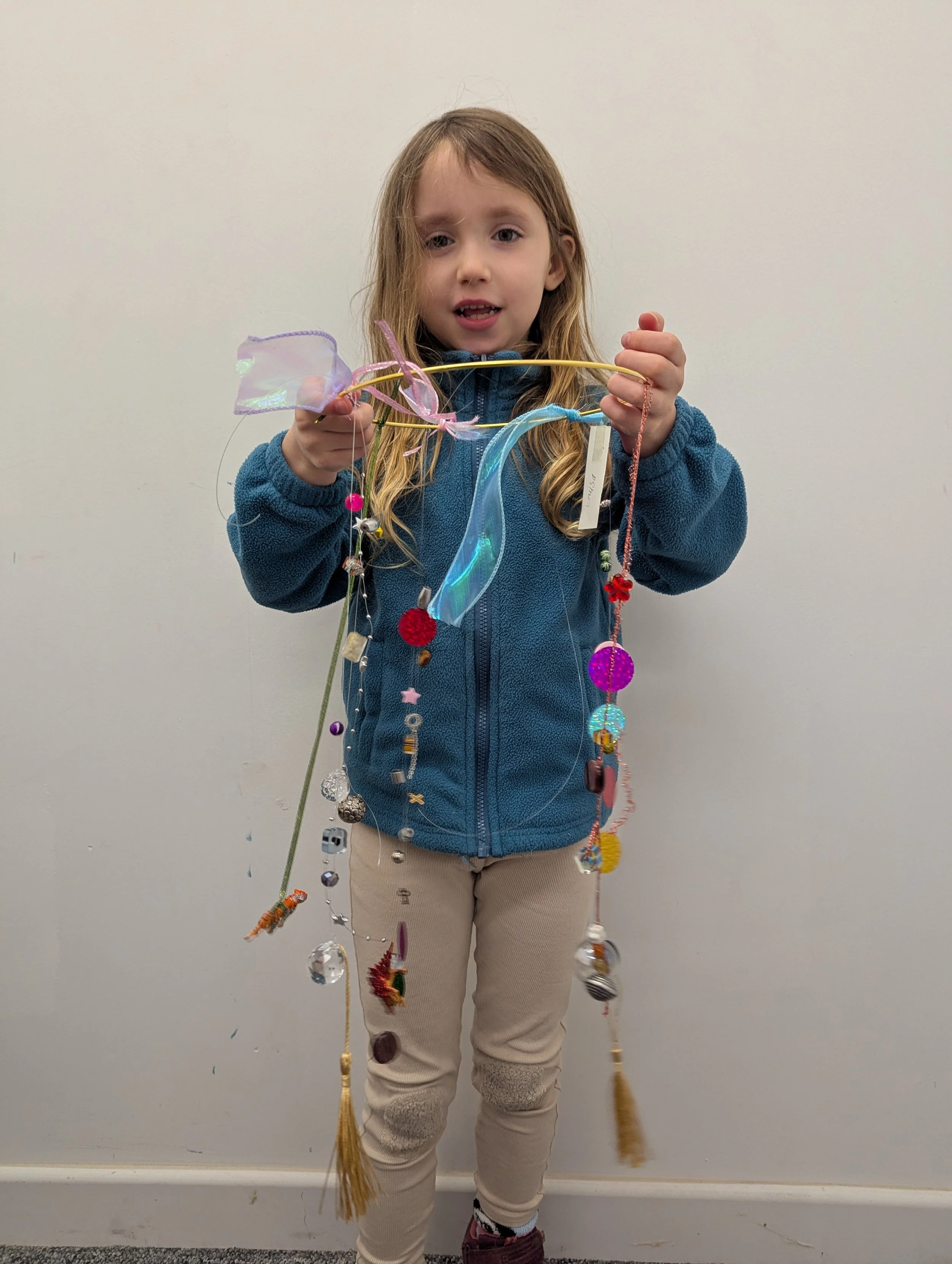 Young girl wearing a blue jacket and beige pants holding a decorated stick with various craft items hanging from it, standing against a plain white wall.