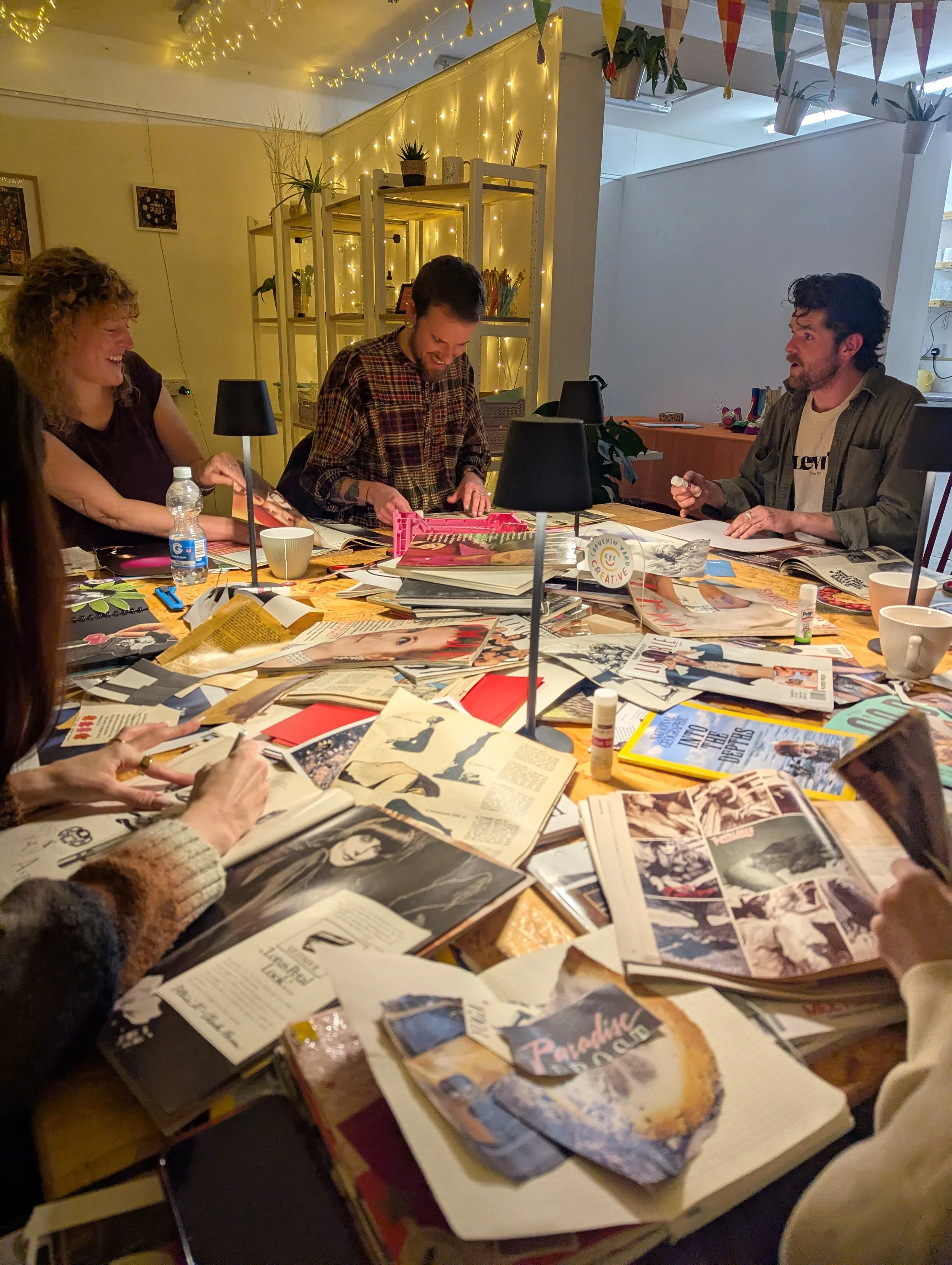 People sitting around a cluttered table with books, magazines, and art materials, engaged in a creative or discussion activity in a warmly lit room with fairy lights and decorative bunting.