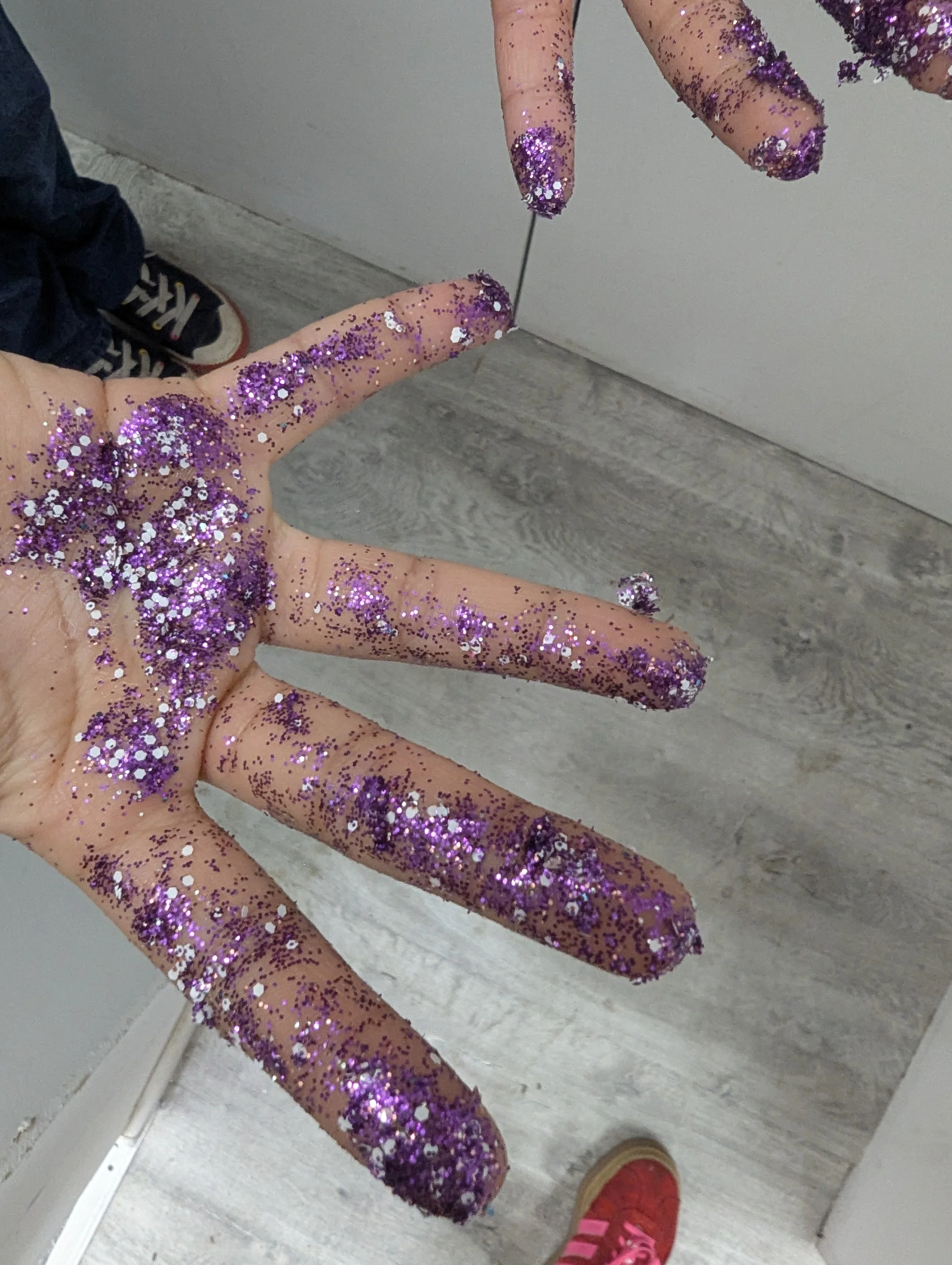 Hand covered in purple glitter, with fingers extended, near a mirror, with a background of wood floor and a wall.