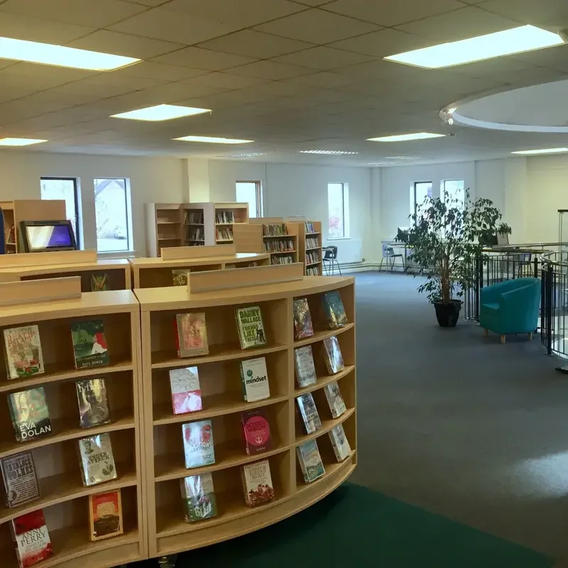 Interior of a library with bookshelves, a large potted plant, and seating areas near windows.