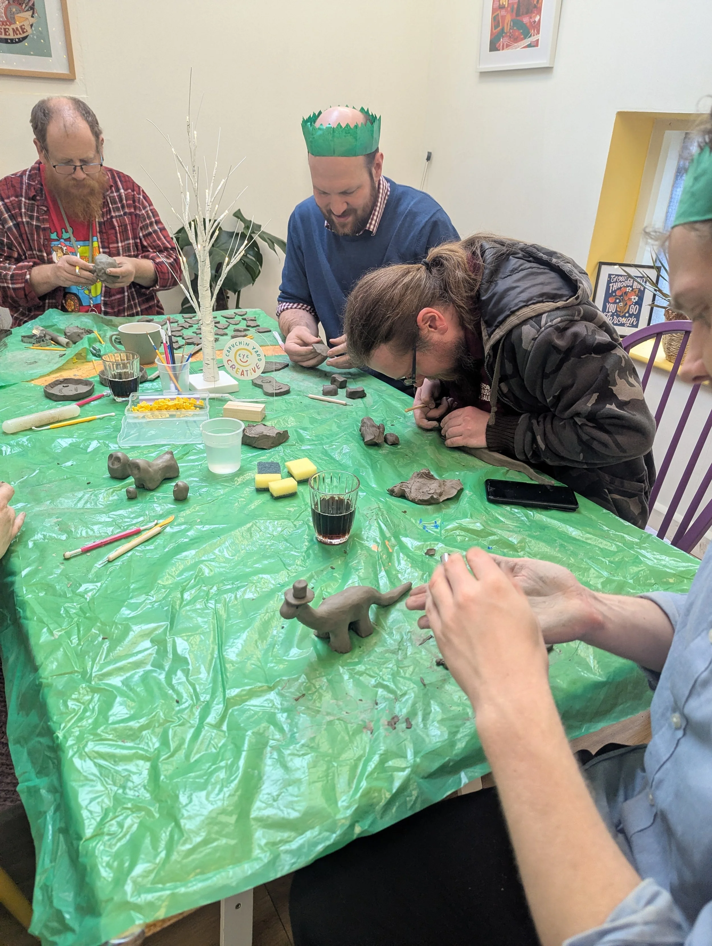 People participating in a clay modeling activity around a table covered with a green plastic tablecloth, decorated with a small white painted tree, a sign that reads 'Crunchin' Creative,' and various clay tools and sculptures.