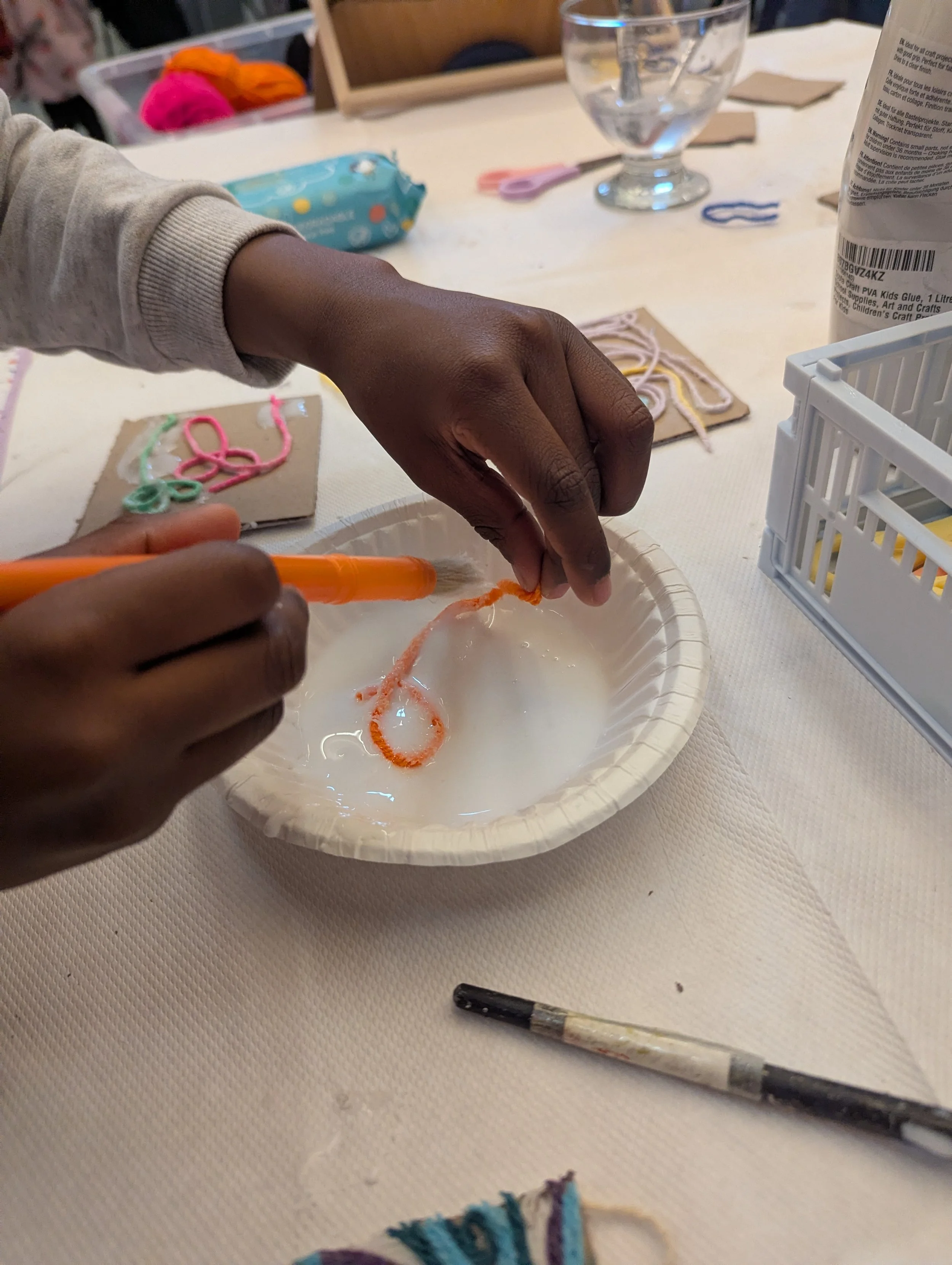 Person making slime with orange pipe cleaner in a bowl of glue and water, surrounded by craft supplies on a table.
