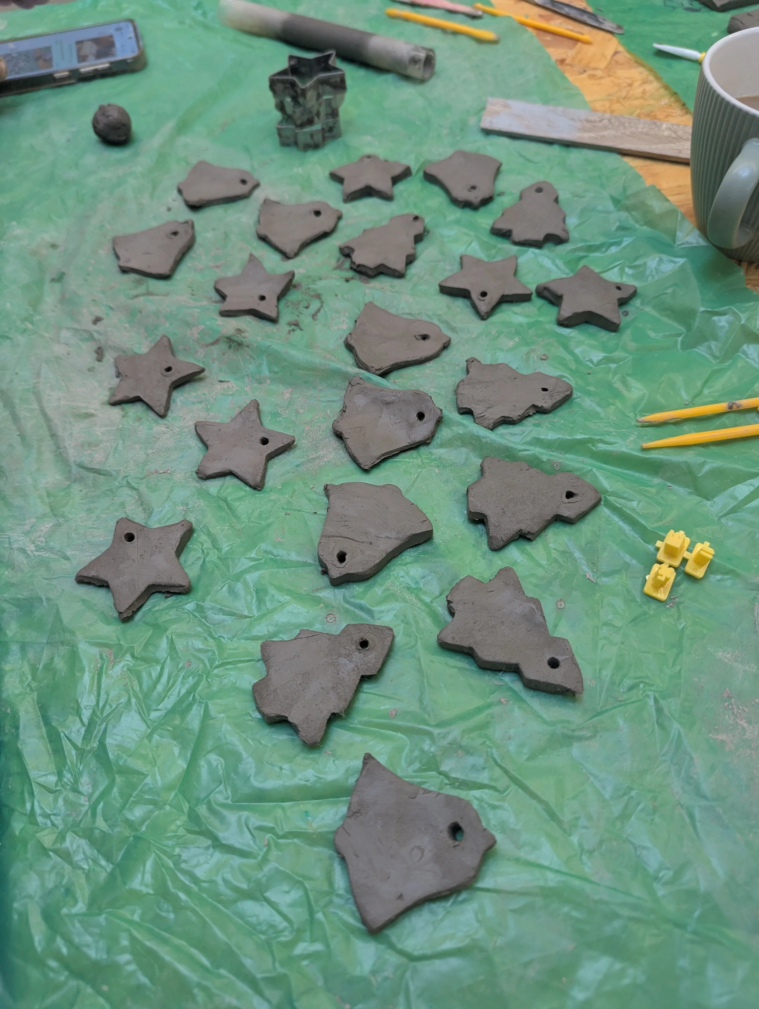 Unfinished gray ceramic ornaments in the shapes of stars, hearts, and trees laid out on a green plastic sheet, with some tools and a cup of coffee nearby.