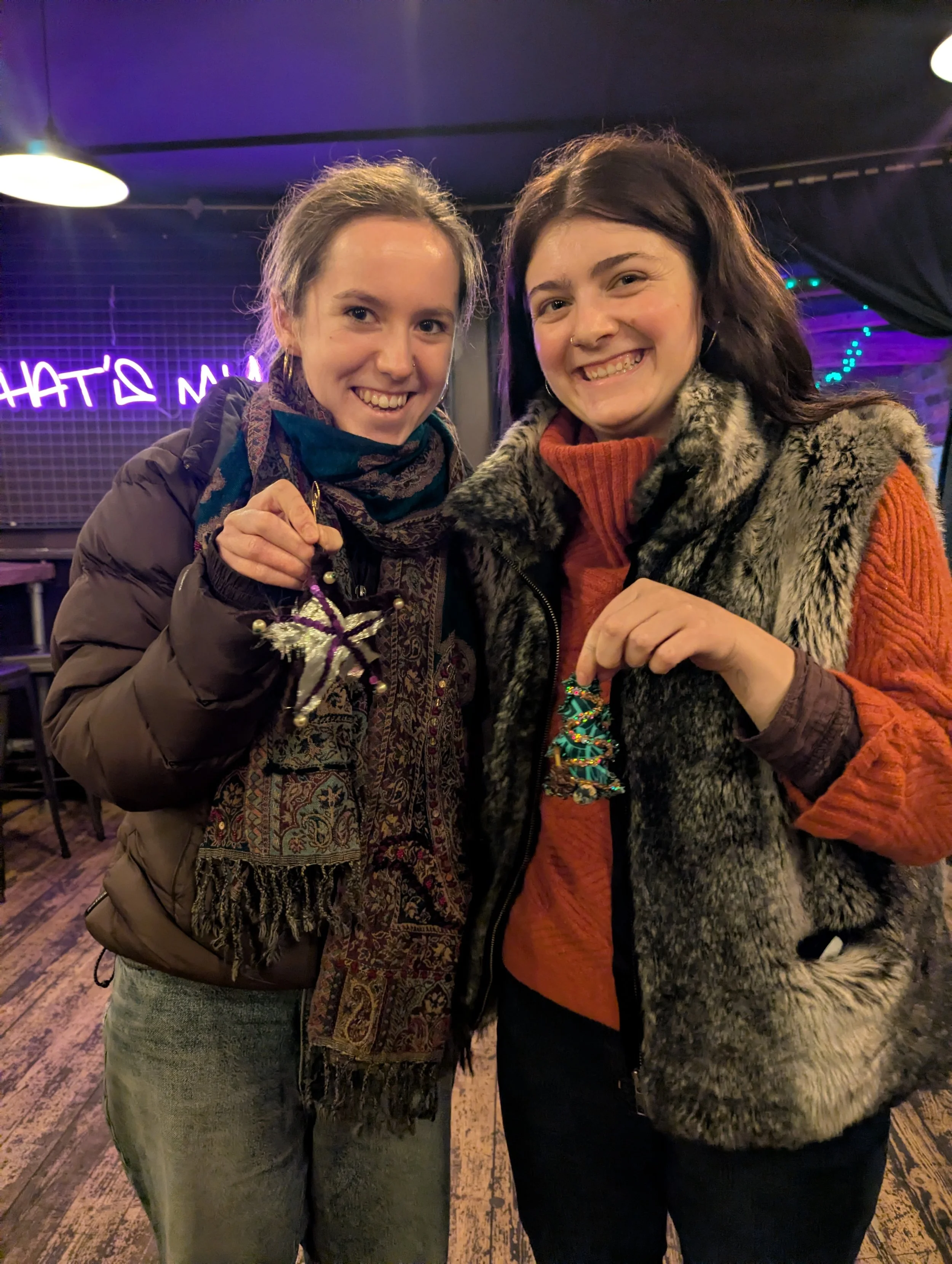 Two women smiling and holding holiday ornaments in a dimly lit venue with neon signs in the background.