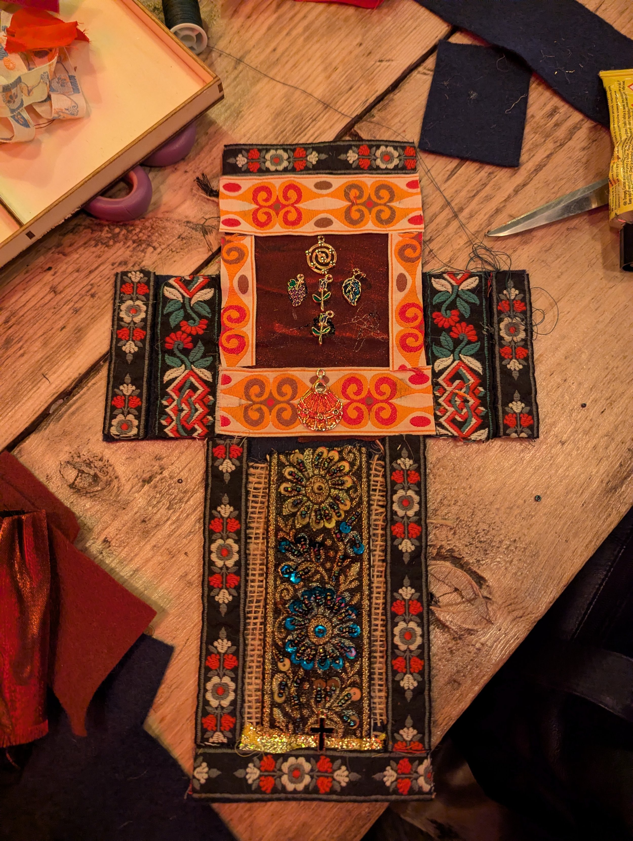 A decorative fabric jewelry box with colorful embroidered patterns, holding various small earrings and pins, placed on a wooden table surrounded by miscellaneous items.