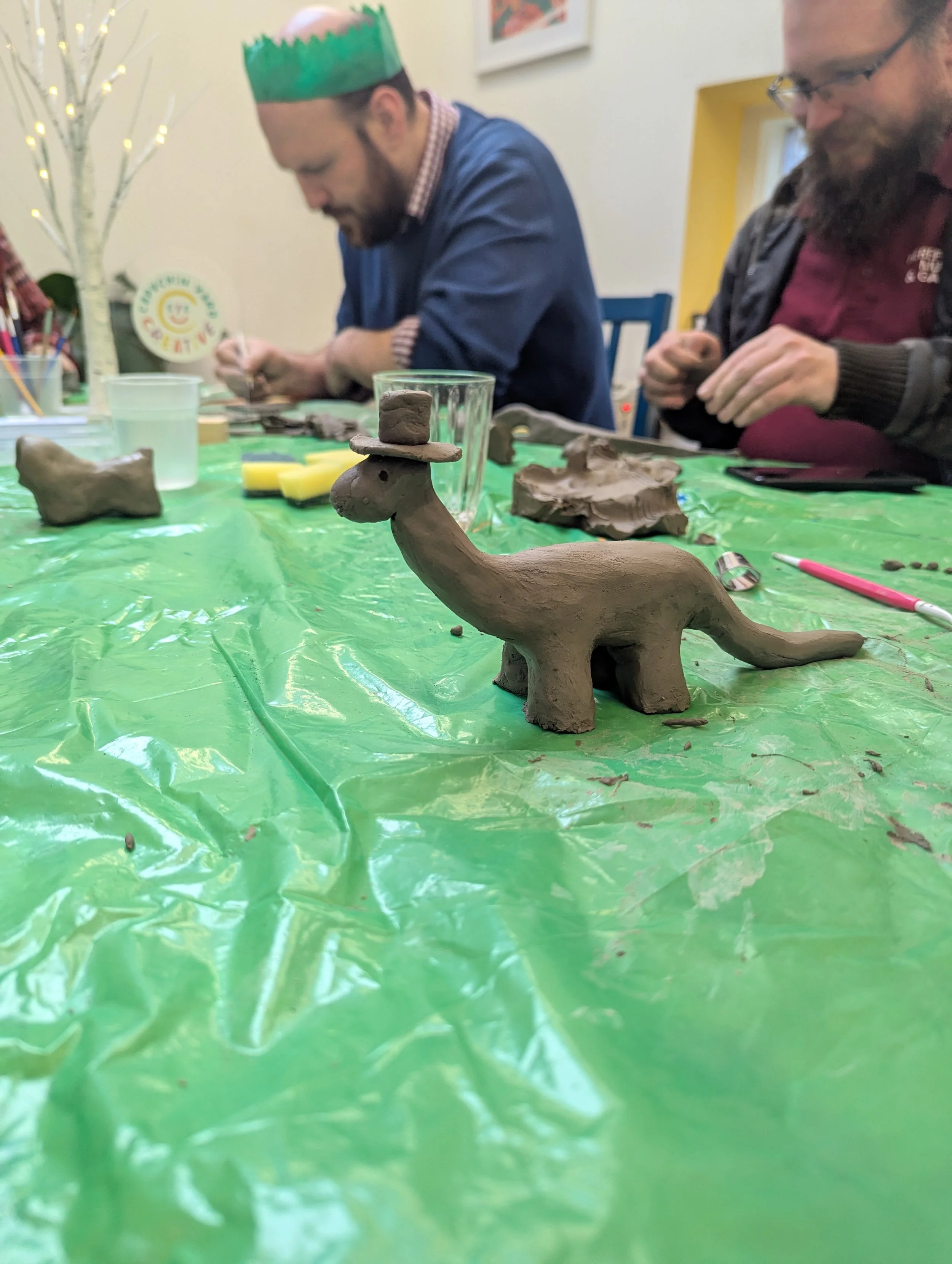 Clay model of a dinosaur with a hat on its head, displayed on a green plastic tablecloth during a pottery workshop, with two people sculpting in the background.