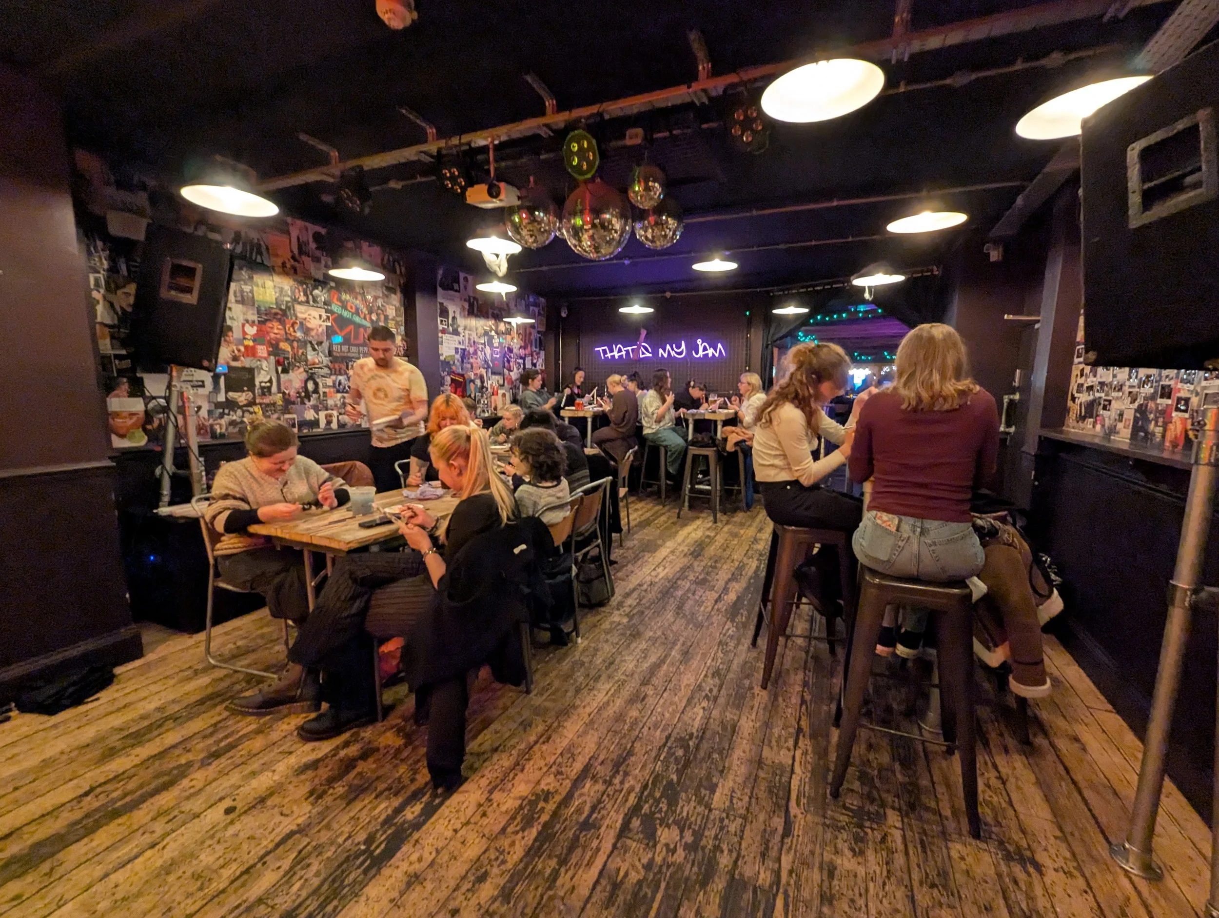 People sitting at tables and bar stools in a dimly lit music venue or bar with a wall decorated with posters and a neon sign reading 'THAT'S MY JAM' in the background.