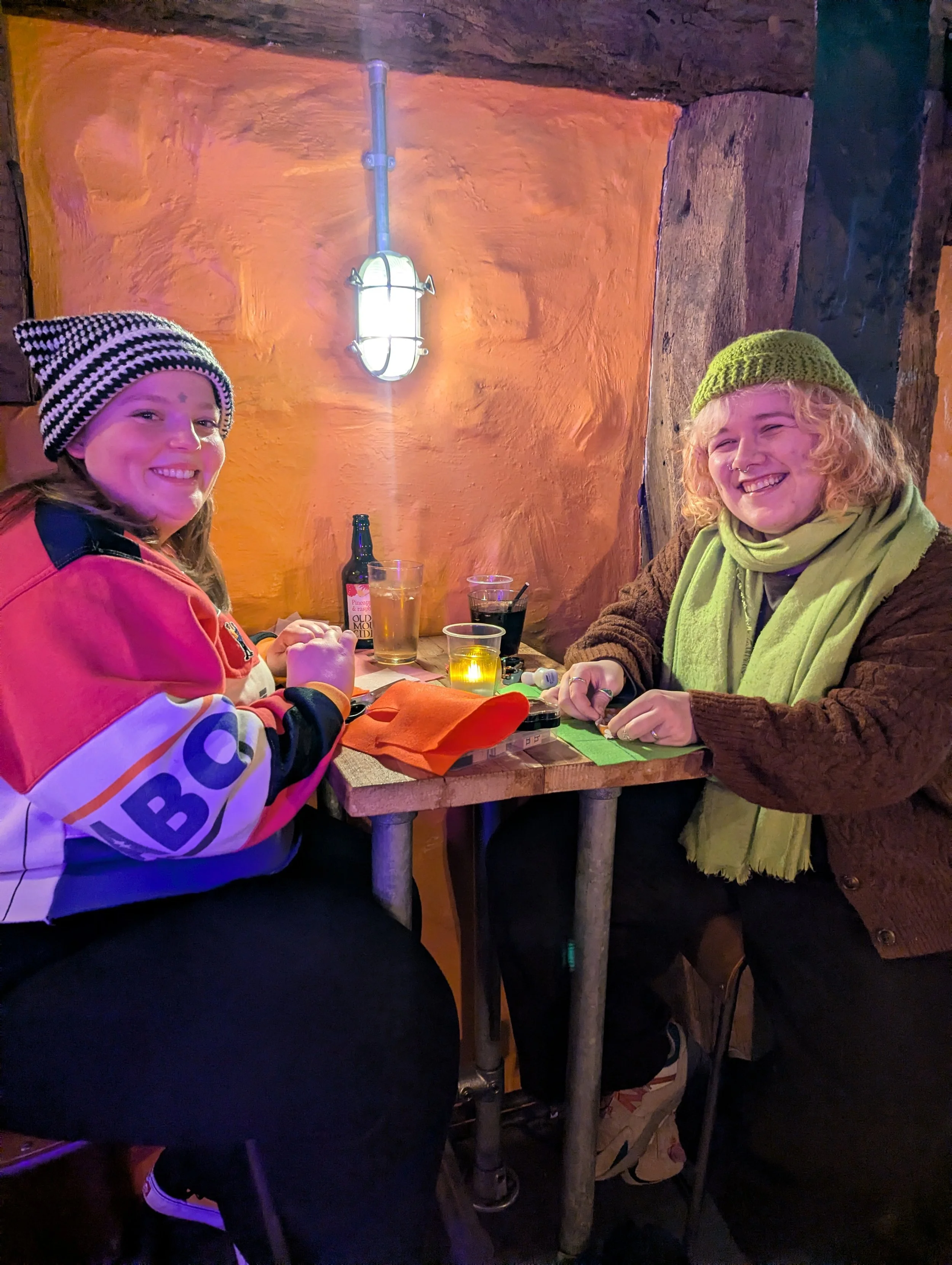 Two women sitting at a wooden table in a cozy restaurant, smiling, wearing winter clothes including hats and scarves, with drinks and snacks on the table, illuminated by warm lighting.