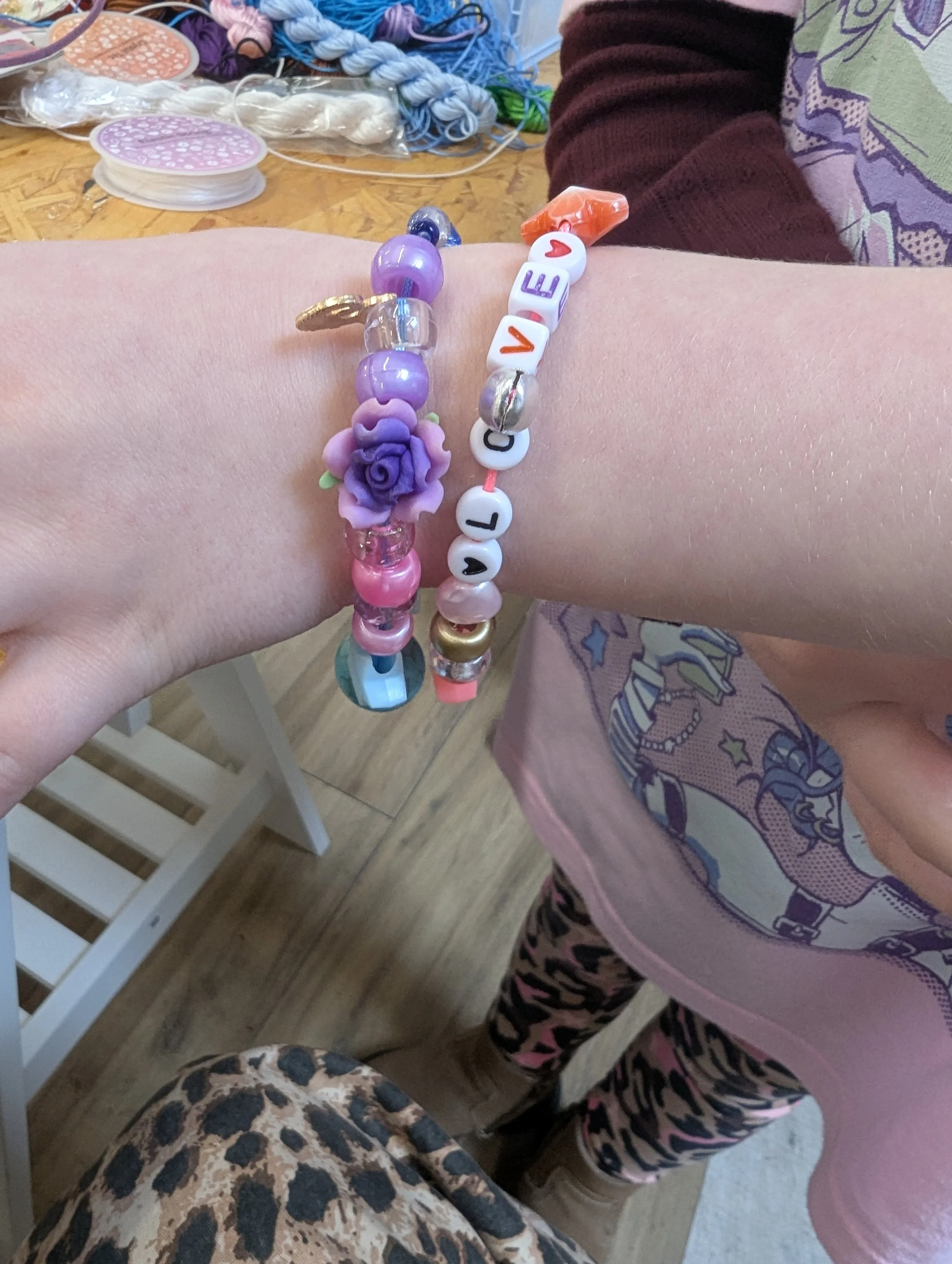 Close-up of a person's wrist with two colorful beaded bracelets, one spelling 'LOVE' and the other featuring a purple flower charm. The background shows crafting materials like thread, beads, and yarn on a wooden surface.
