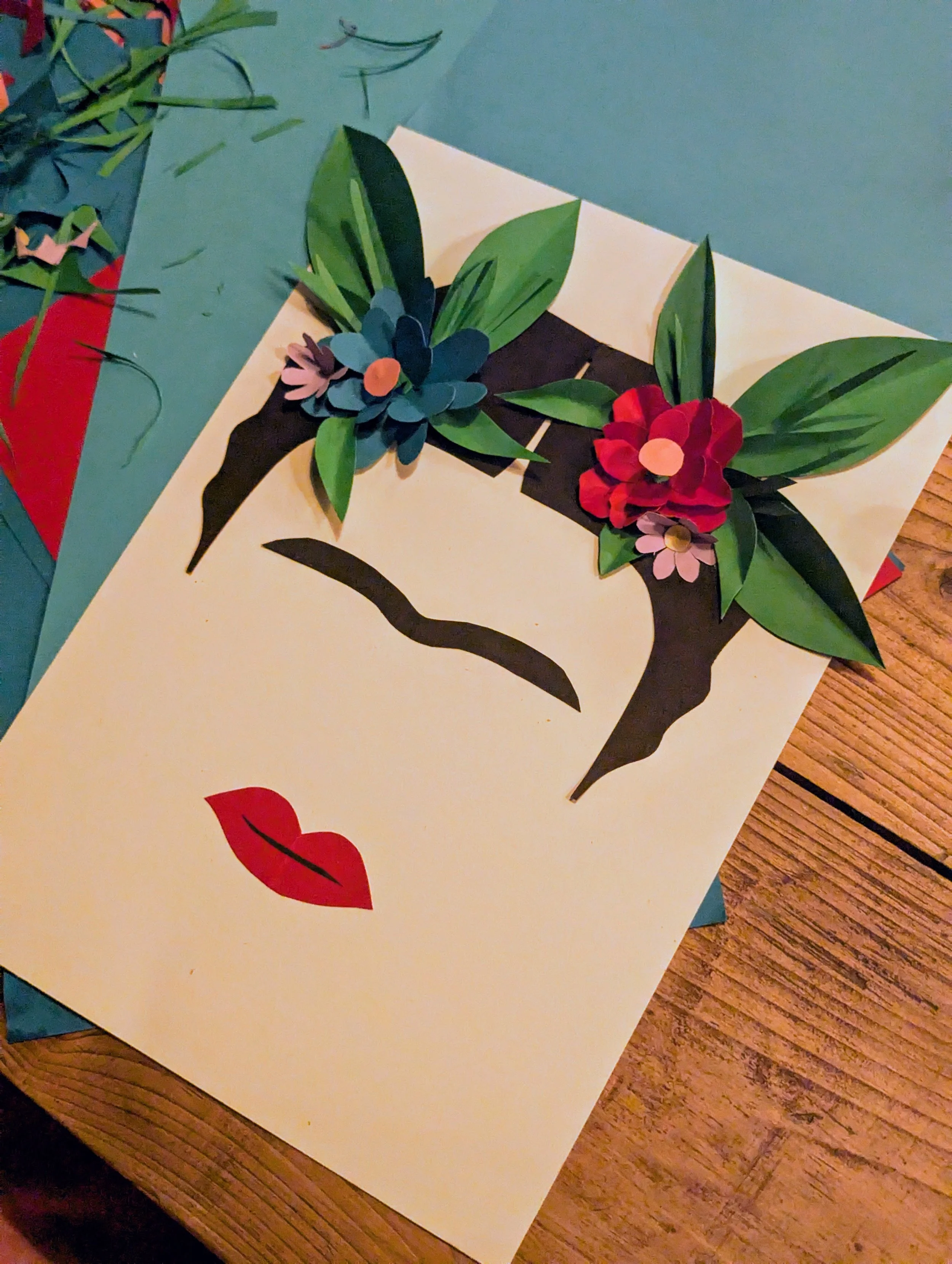 A greeting card with a face made of paper cutouts. The face has red lips, closed eyes, and has attached paper flowers and leaves on top of the head like Frida Kahlo.