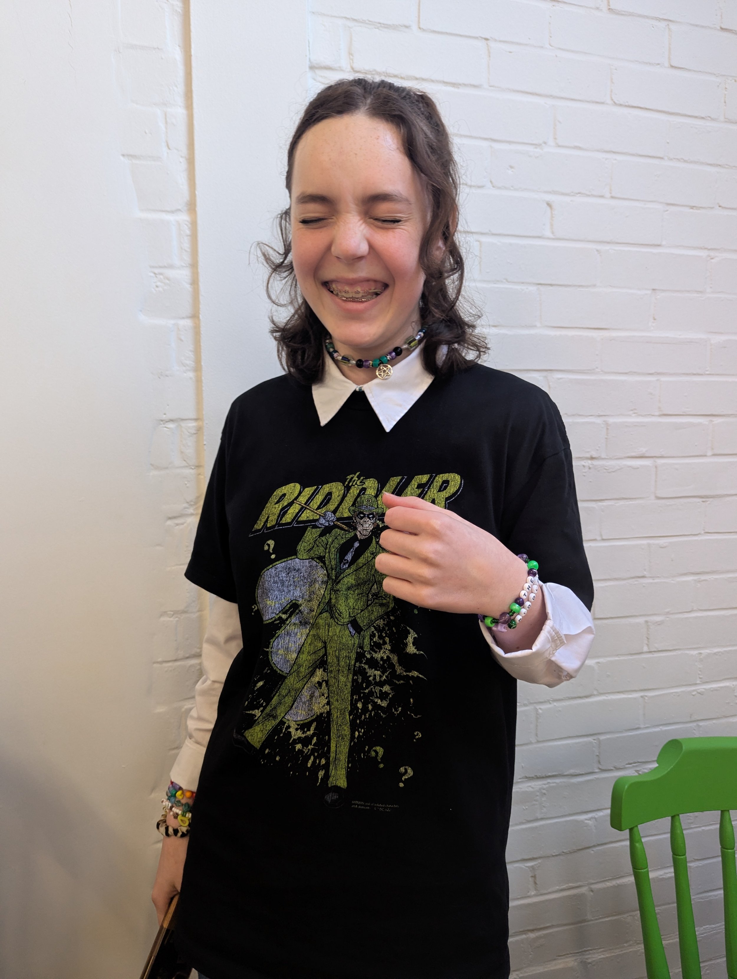 Young girl with braces laughing, wearing a black 'Rider' T-shirt over a white shirt, with colorful jewelry, standing in front of a white brick wall.