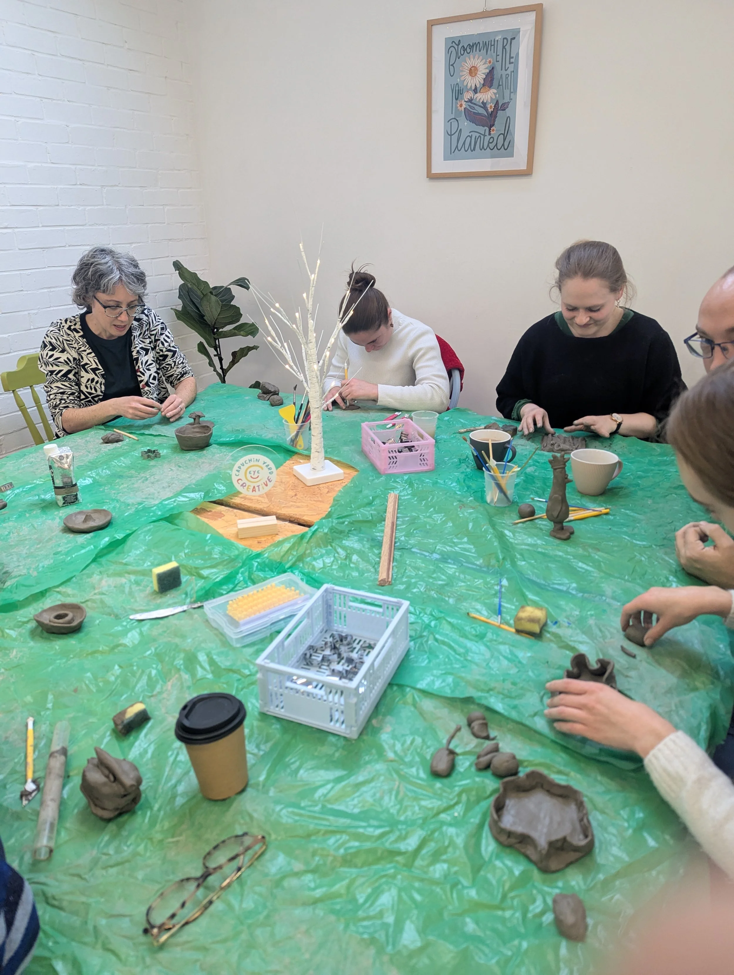 People participating in a ceramic art workshop, working on clay projects around a green plastic-covered table, with tools and pottery supplies visible.