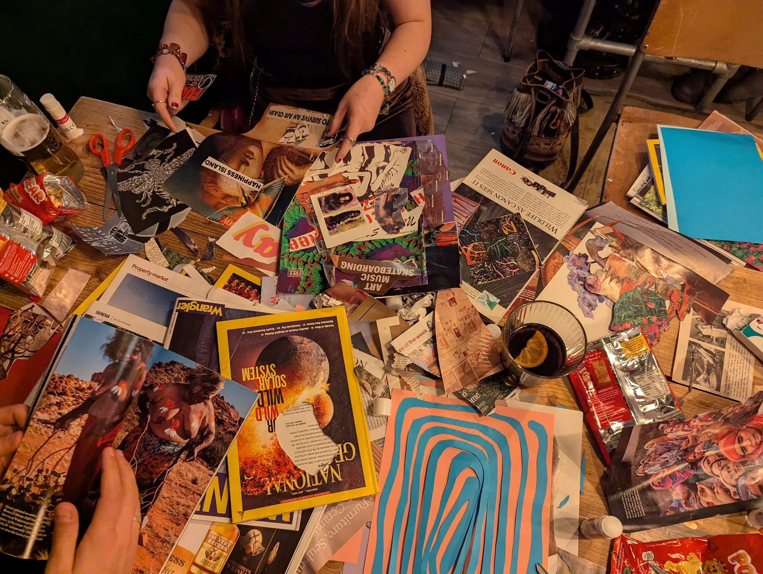 A cluttered table with magazines, photographs, snacks, a glass of soda with lemon, and a person's hands looking through the magazines.