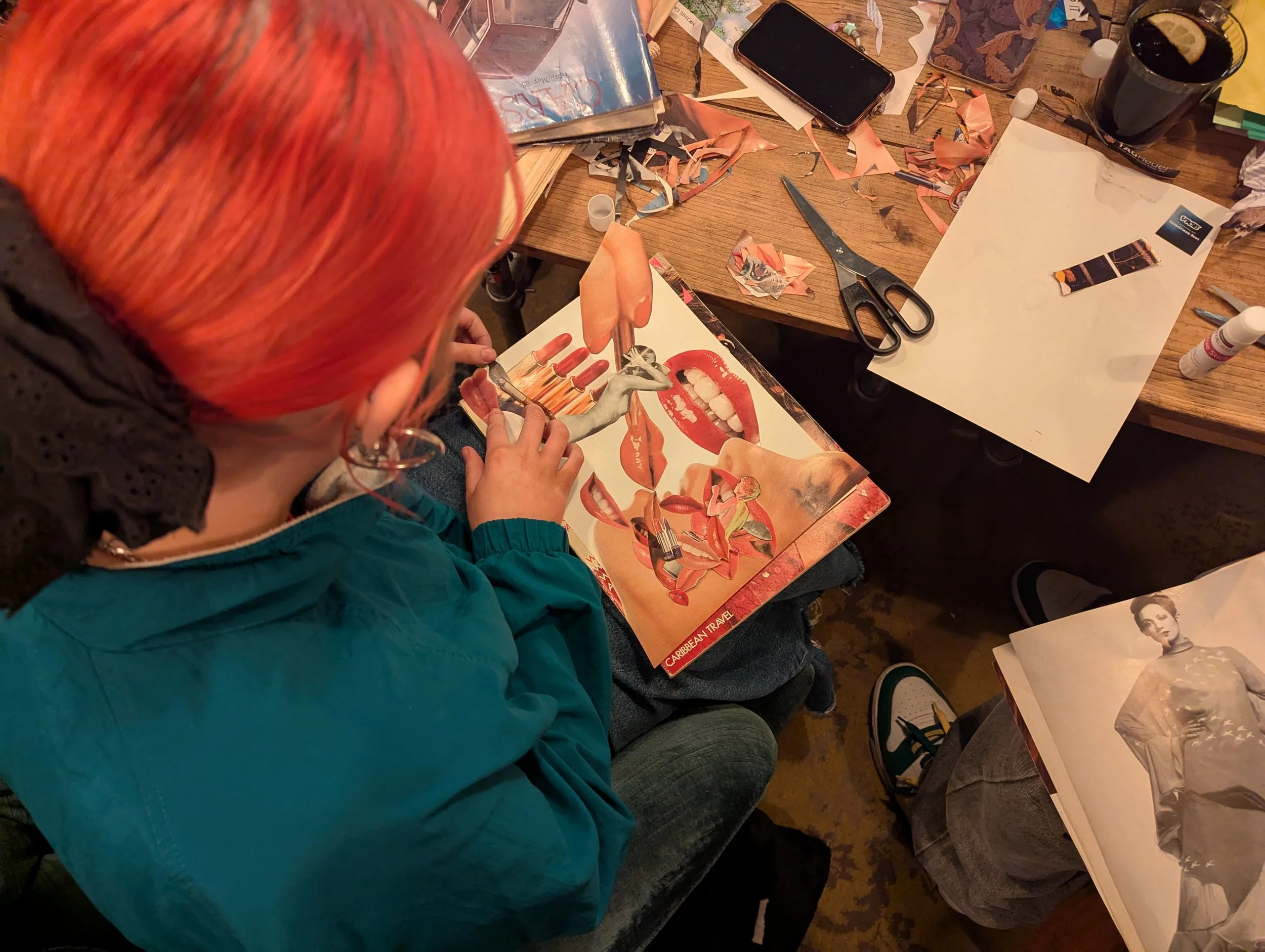 A person with bright orange hair sitting at a cluttered table, working on a collage with cut-out magazine images of lips, a woman, and various objects. The table has scissors, glue, a white sheet of paper, and other crafting materials.