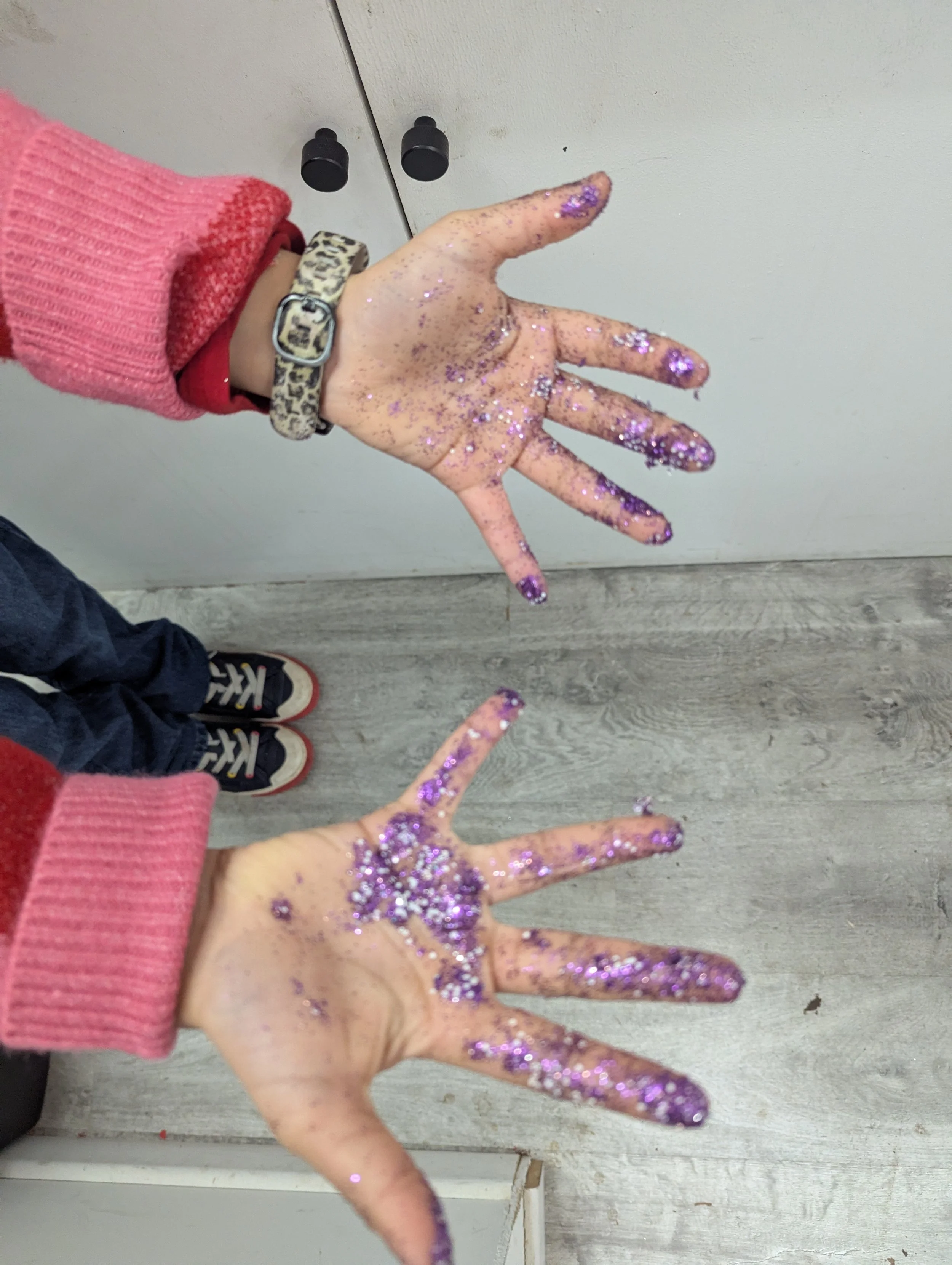 Hands with purple glitter on fingers and palms, wearing pink and red sweaters, and a leopard print watch. Floor is light-colored wood, background is a white cabinet.
