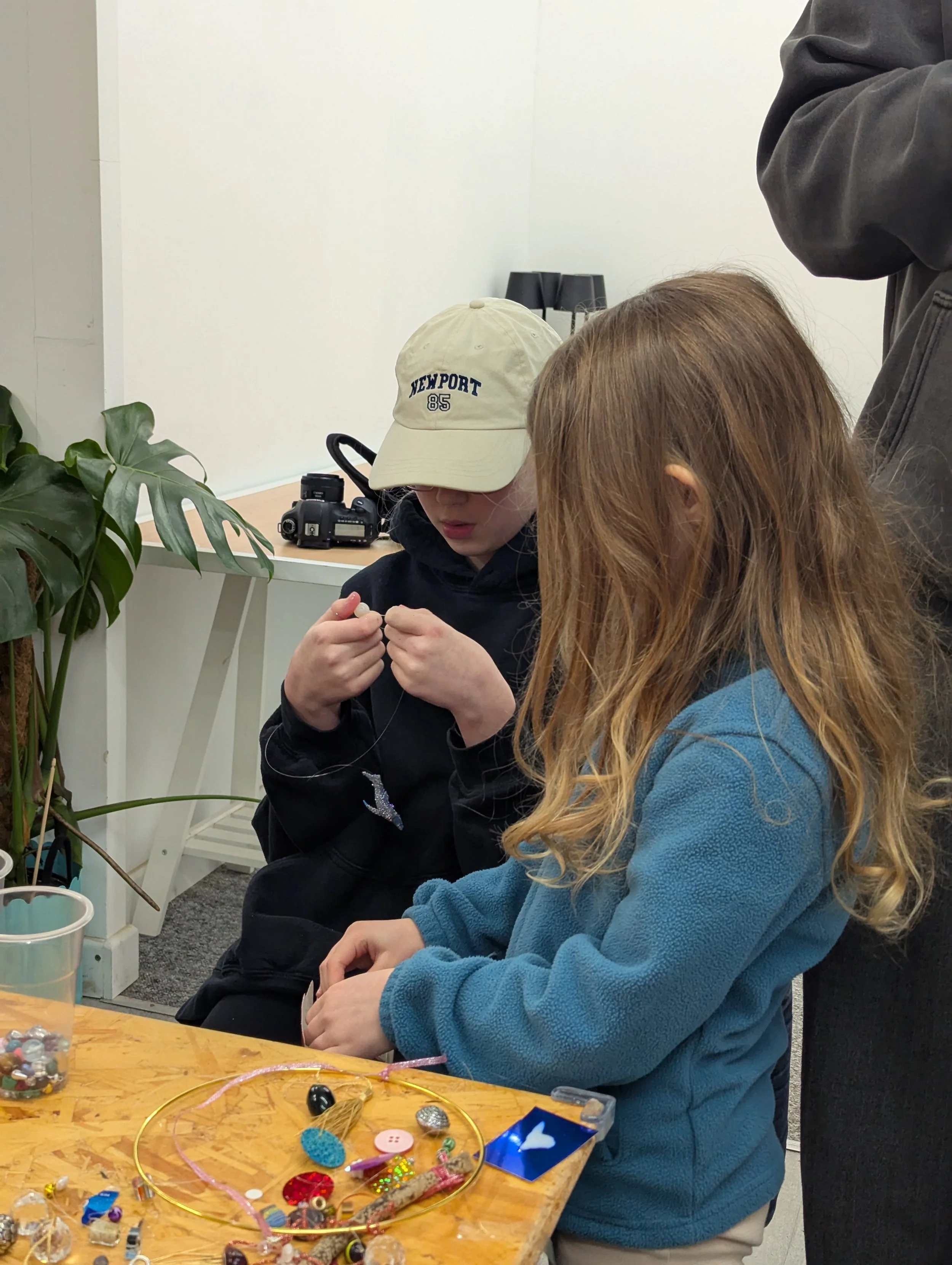 Two children with long hair, one wearing a beige cap and black hoodie, and the other with red hair and a blue jacket, sitting at a table with various craft supplies like beads and buttons, working on a craft project. An adult standing nearby, and a c