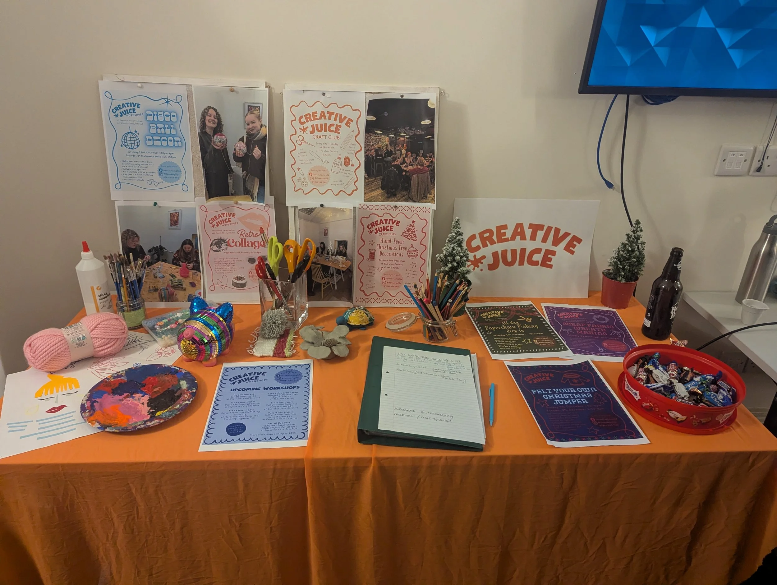 Table with craft supplies, notices, and decorations at a creative juice event, including yarn, art projects, scissors, and promotional materials.