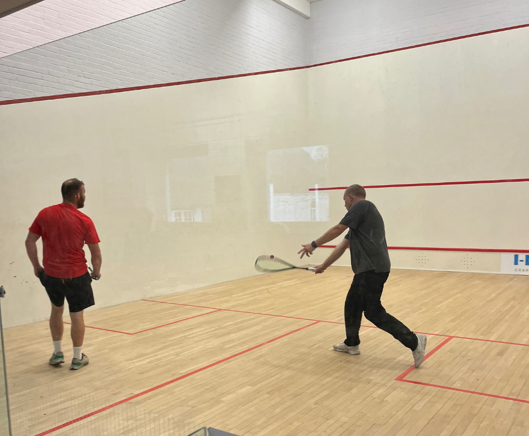 Two men playing squash in an indoor court, one about to hit the ball with a racket while the other observes.