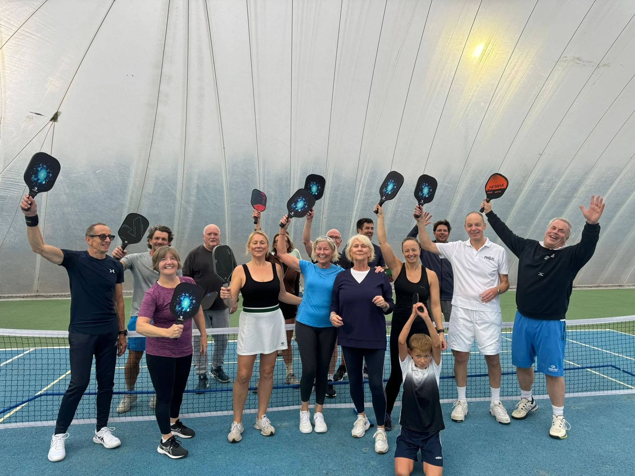 Group of twelve people on an indoor tennis court holding paddle rackets, smiling, celebrating, and raising their arms, with a young boy in the foreground holding a paddle above his head.