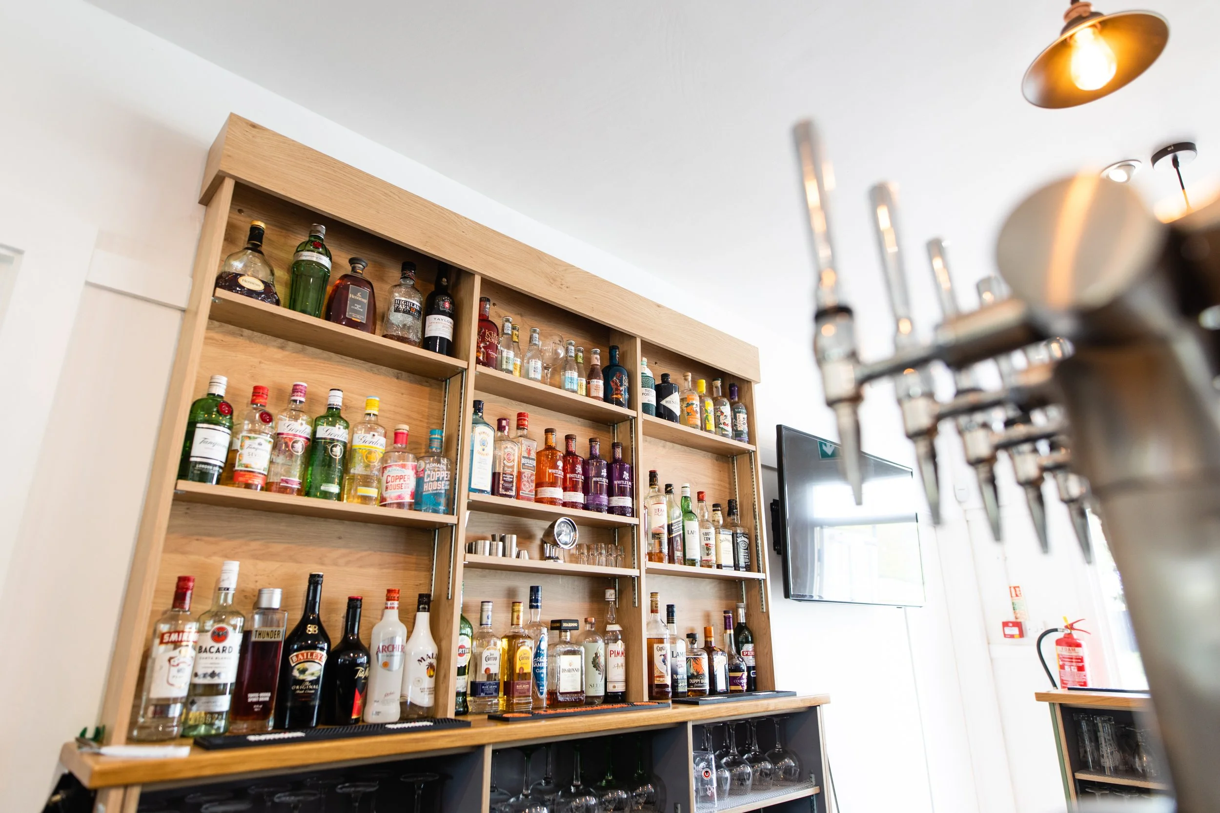 A wooden bar shelf filled with various bottles of liquor, with a tap system in the foreground and a wall-mounted TV on the right.