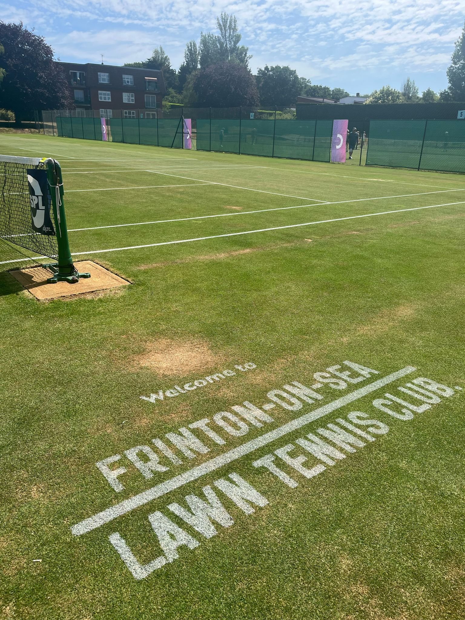 A tennis court with the words 'Welcome to FRUITLOOT.NETLAND TENNIS CLUB' painted in white on the grass near the court entrance.