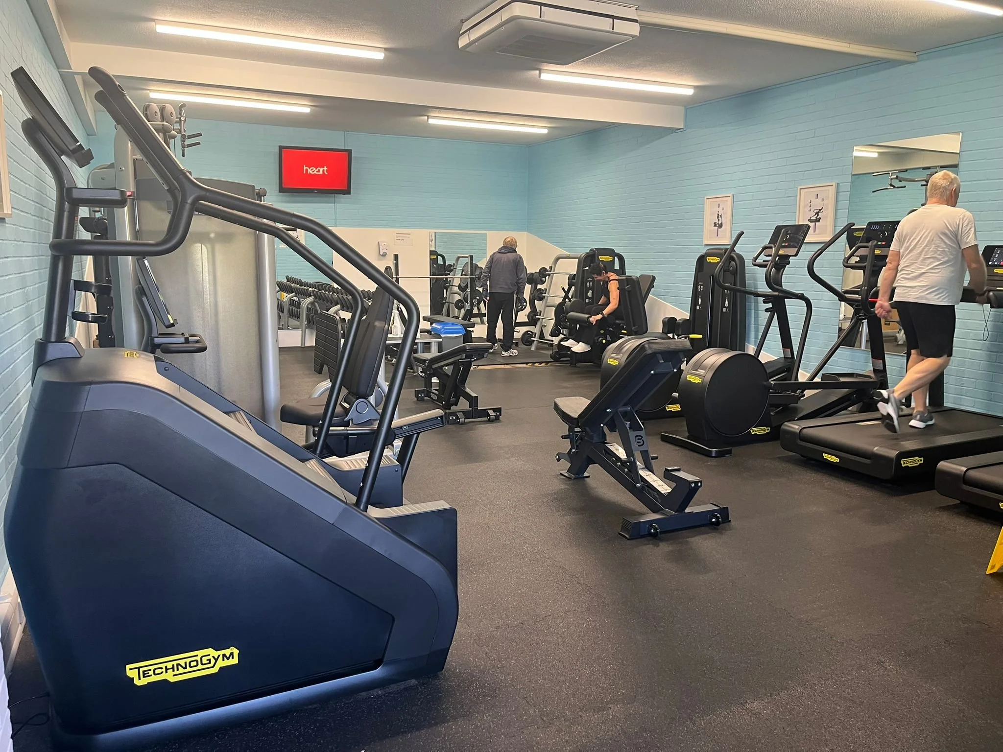 Indoor gym with various exercise machines, including treadmills, ellipticals, and weight machines, with a few people working out and a red digital display that says 'heart' on a blue painted wall.
