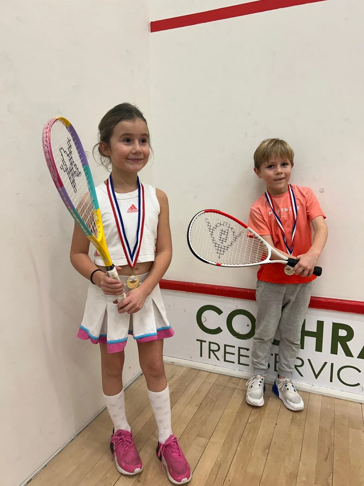 Two children, a girl and a boy, standing indoors with tennis rackets and medals around their necks, smiling.
