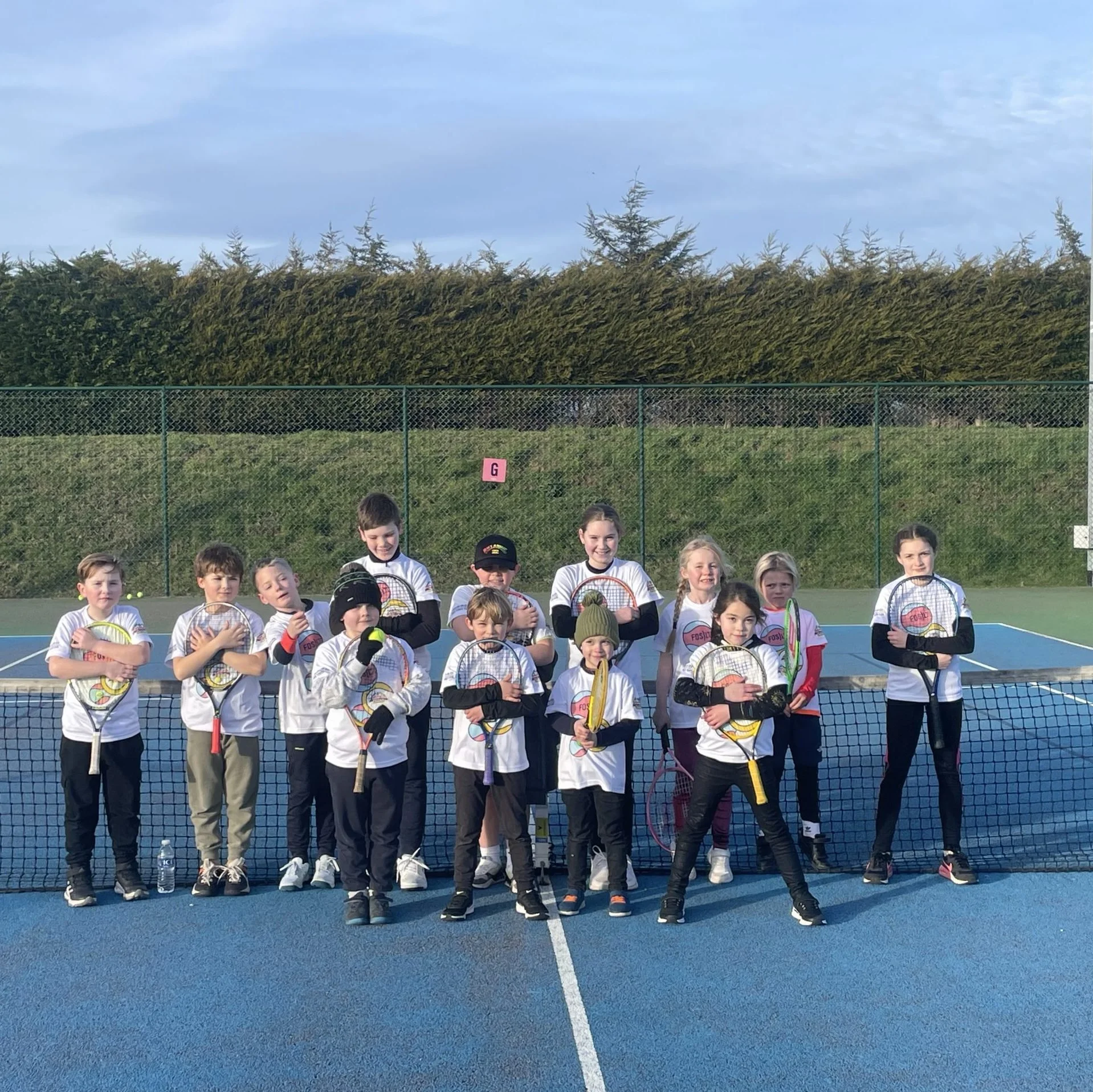 Group of children on a tennis court, holding tennis rackets and wearing sports uniforms, posing together for a photo.