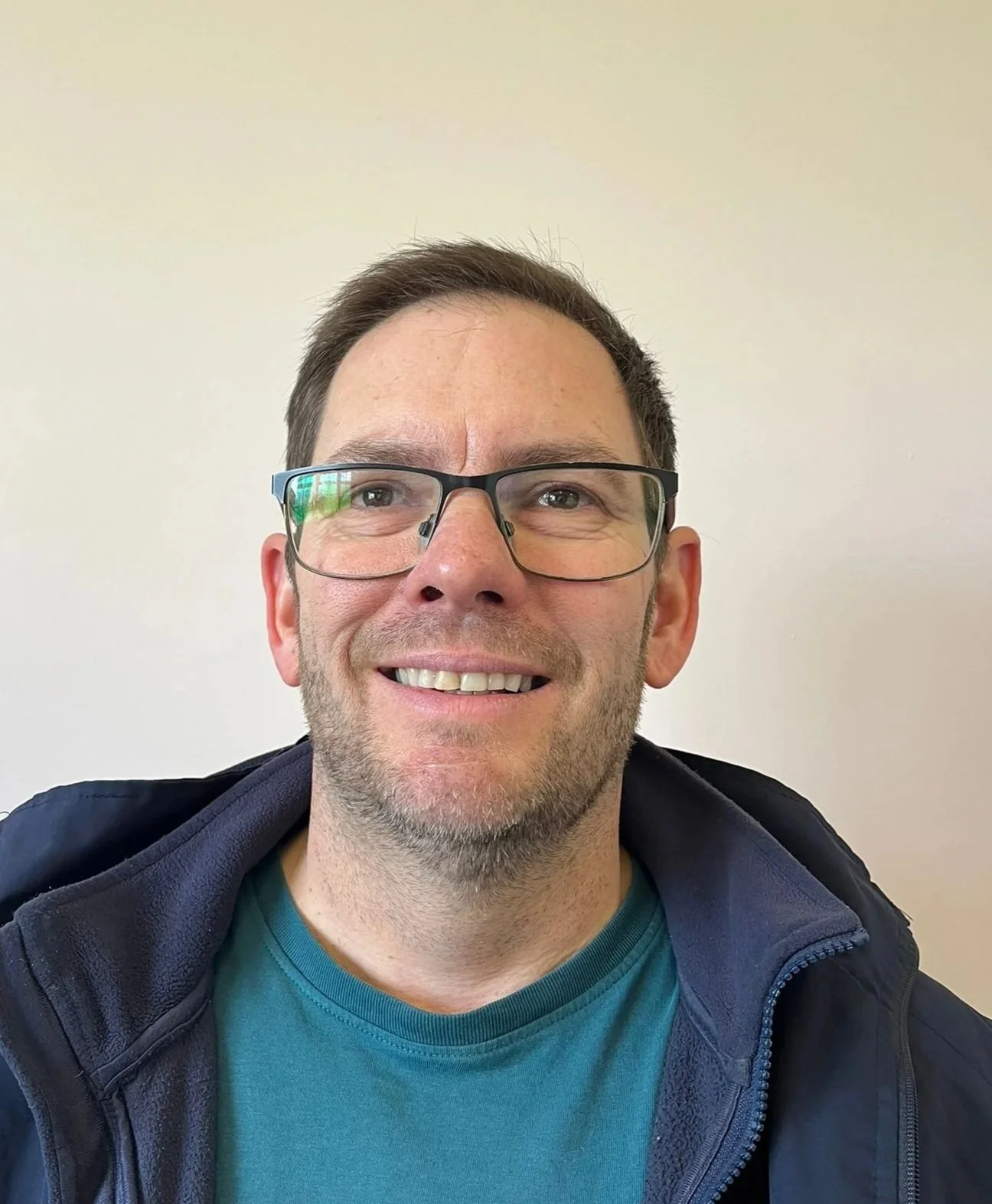 A smiling man with glasses and short dark hair, wearing a blue shirt and navy jacket, against a light beige background.