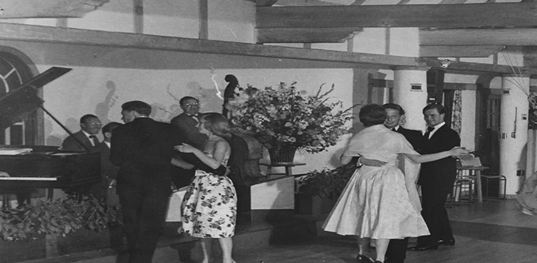 Couples dancing in a vintage indoor setting with a large floral arrangement in the background, a piano, and a decorated stage area.
