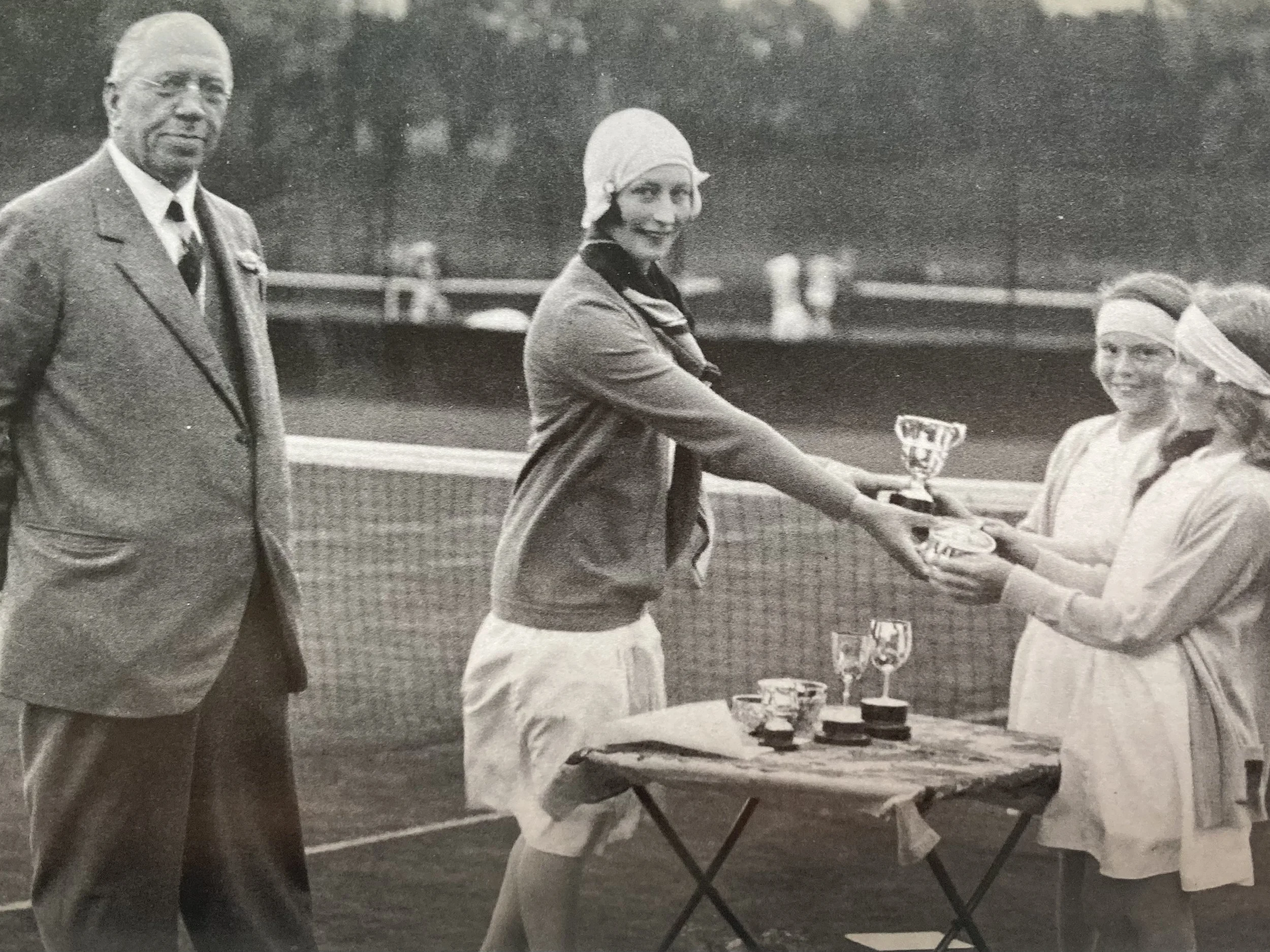 A black and white photo of a woman in a tennis outfit receiving a trophy from another woman, with a man standing nearby, on a tennis court.