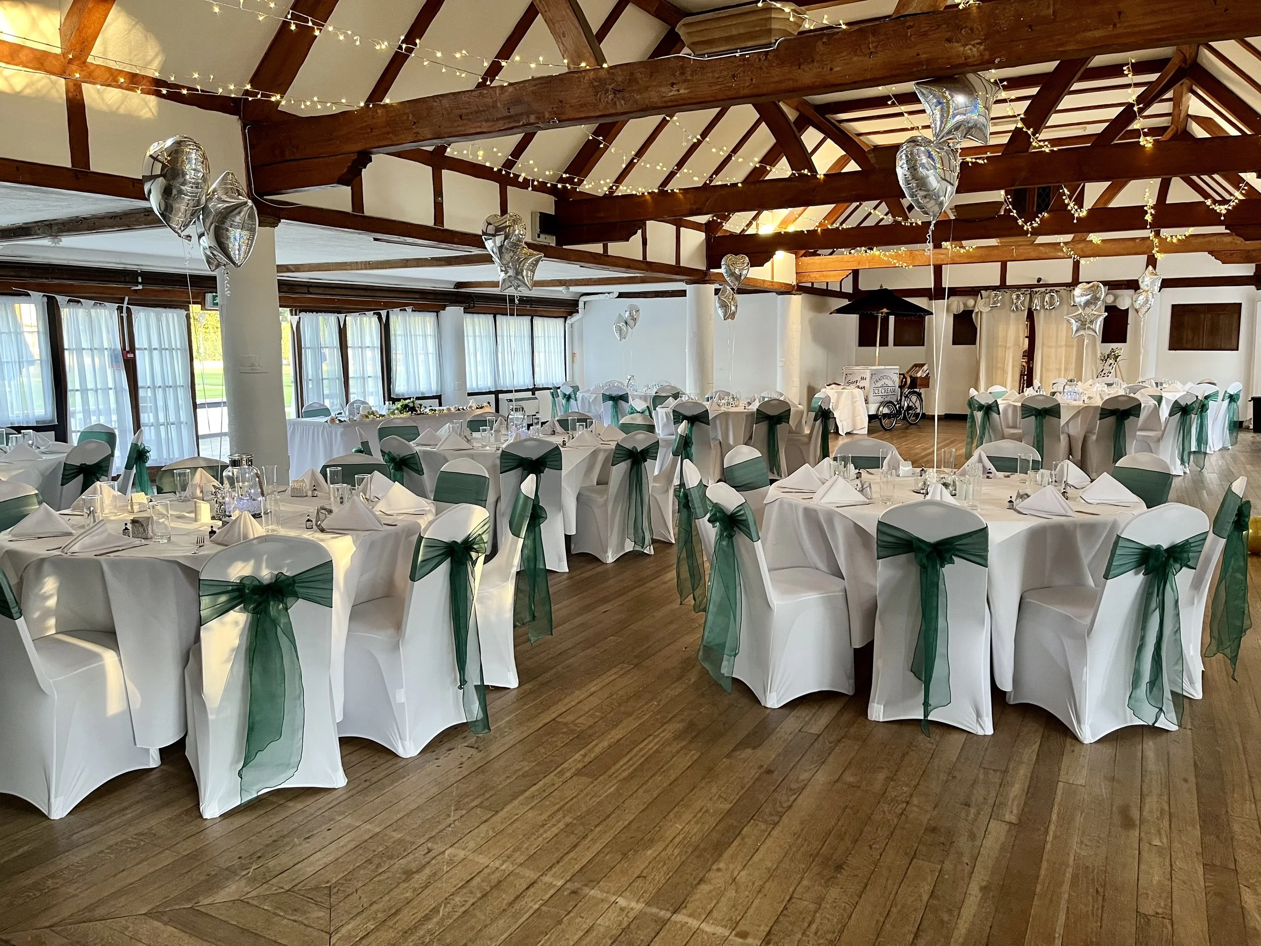 A beautifully decorated event hall with round tables covered in white tablecloths, green chair sashes, and place settings including napkins, glasses, and small black bottles. Silver heart-shaped balloons are hanging from the ceiling, and string lights are draped across the wooden beams of the ceiling. The windows are large with sheer curtains, and there is natural light coming in.